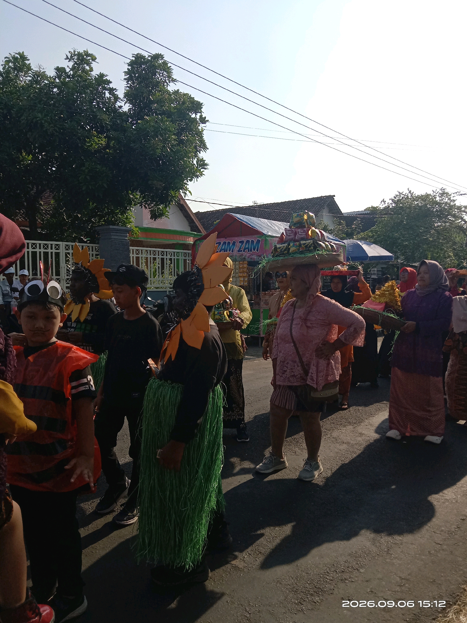 A group of people are participating in a parade in Sepanyul, Indonesia. The parade appears to be a cultural or festive event, with participants dressed in traditional or costume attire. In the foreground, a young boy is wearing an orange and black striped costume with large googly eyes and a fake mustache. Beside him, another person is wearing a green grassy skirt. Further back, a woman is carrying a large stack of items on her head, possibly food or offerings. Other individuals are also visible, some carrying baskets or wearing colorful clothing and headscarves. The background shows houses and trees, suggesting a street in a residential area. Power lines are visible across the sky. The image is dated "2026.09.06 15:12".