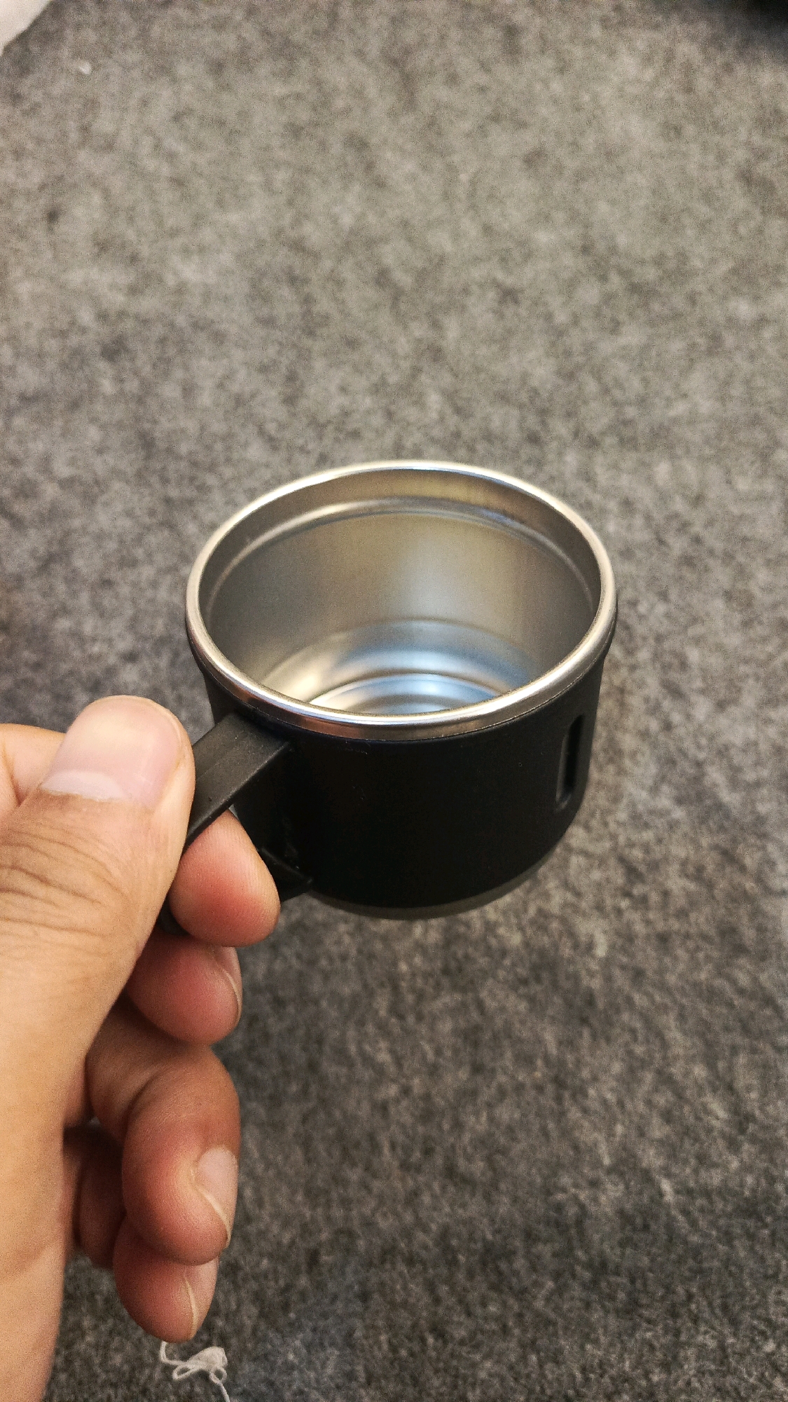 A close-up shot shows a person's hand holding a small, black mug with a silver interior. The mug has a simple handle and appears to be made of metal. The background is a textured, grey surface, possibly a carpet or fabric. The image is well-lit, with the focus clearly on the mug and the hand. The location is specified as Babadan, Indonesia, though this is not visually evident in the image itself.