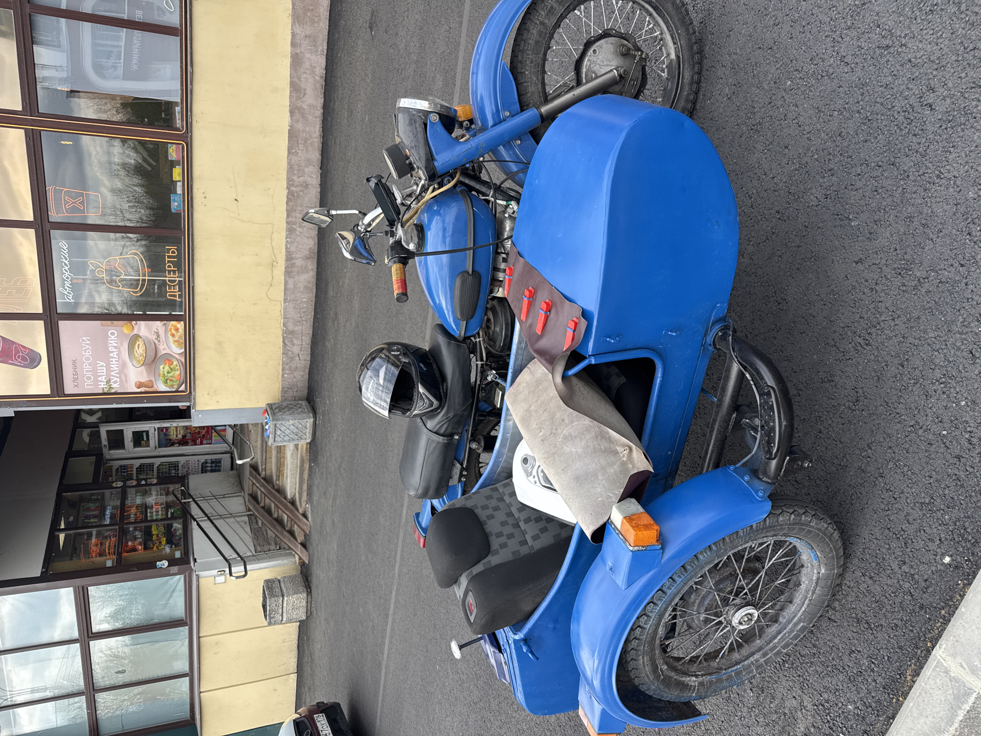 A blue motorcycle with a sidecar is parked on an asphalt surface next to a yellow building in Saint Petersburg, Russia. The sidecar contains a black helmet, a seat with a checkered pattern, and a crumpled beige cloth. The building has a storefront with windows displaying advertisements for food and drinks. A ladder leans against the steps leading up to the store entrance. In the reflection in the window, a white vehicle is visible.