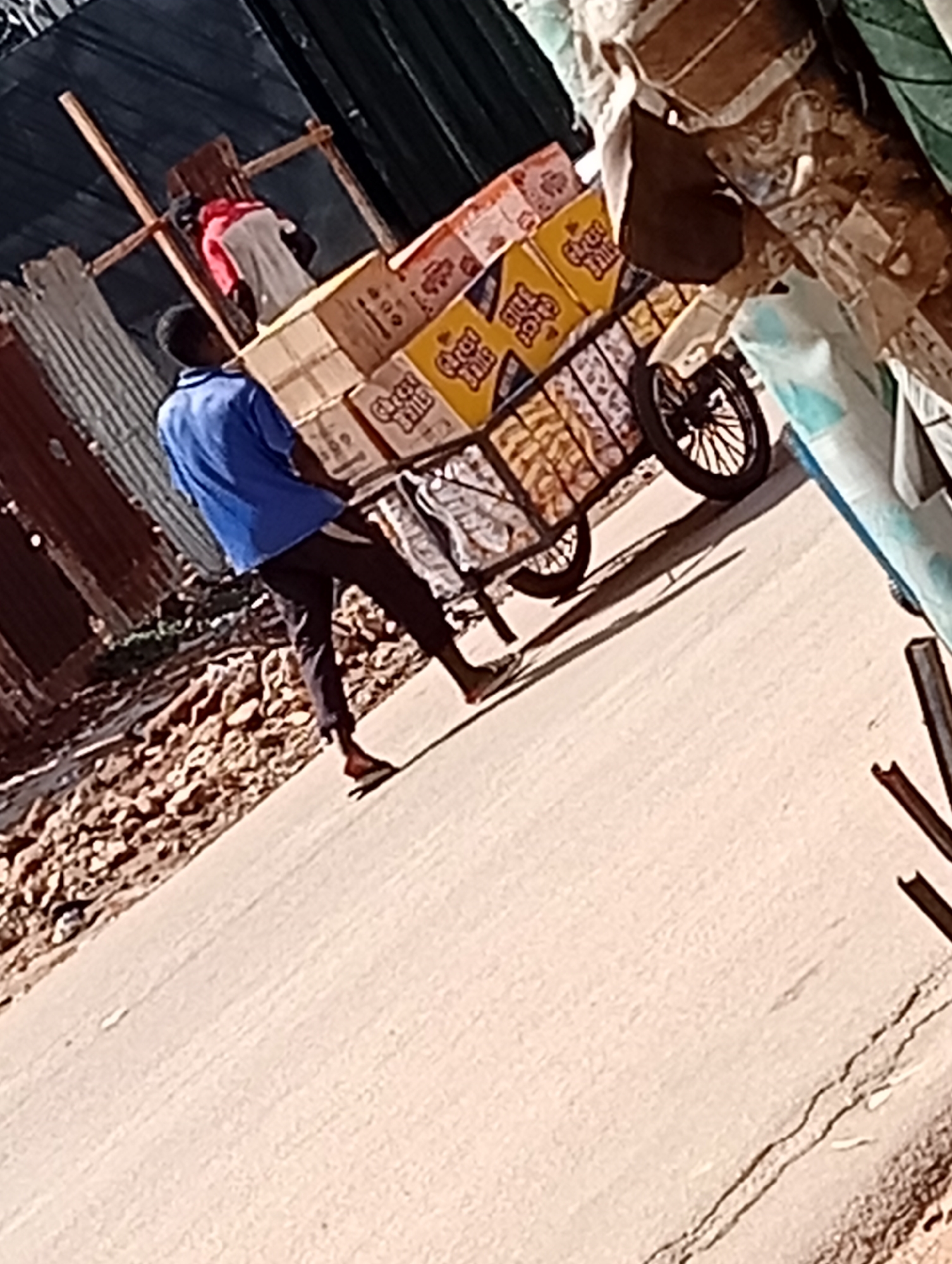 A man in a blue shirt and dark pants pulls a cart loaded with boxes of Great Balls juice down a paved road in Maiburiji, Nigeria. The surroundings include corrugated metal buildings and debris on the roadside.