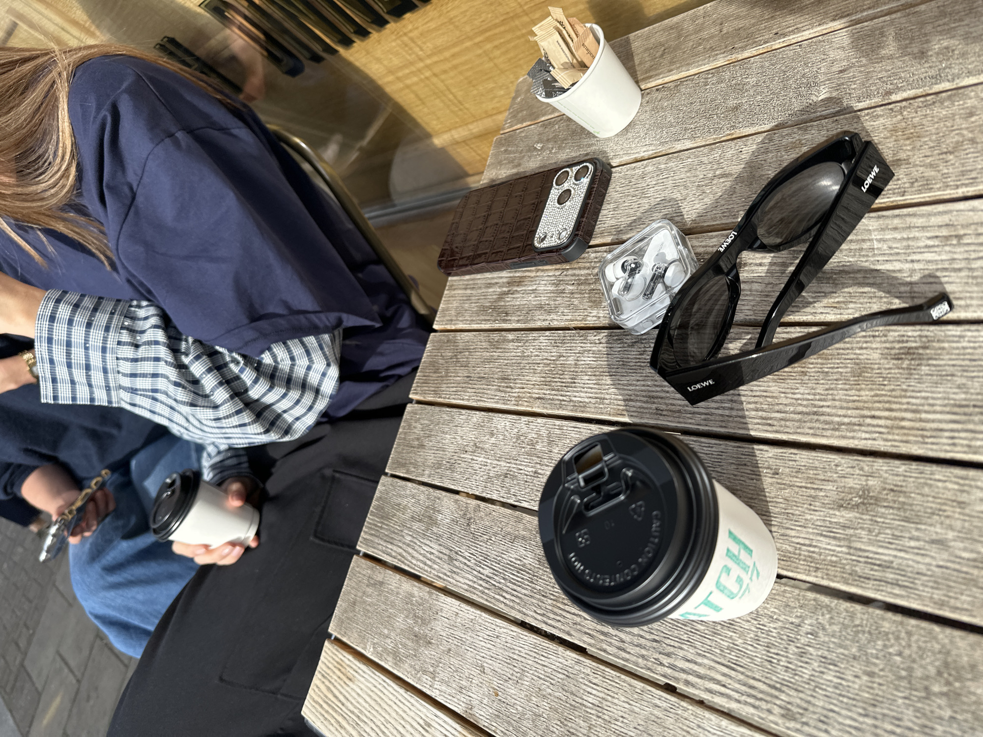 The image captures a casual scene on a wooden outdoor table, likely in London, United Kingdom. A person sits to the left, wearing a navy blue shirt layered over a blue and white checkered long-sleeved shirt, with black pants. They are holding a white paper coffee cup in their hands.
On the table, various items are scattered: a brown phone case with a crocodile-like texture, a transparent case containing white earphones, a pair of black Loewe sunglasses, a white paper cup filled with sugar packets, and another coffee cup with a black lid bearing the name "Watch." The angle suggests a candid snapshot, providing a glimpse into a personal moment at an outdoor cafe or seating area.