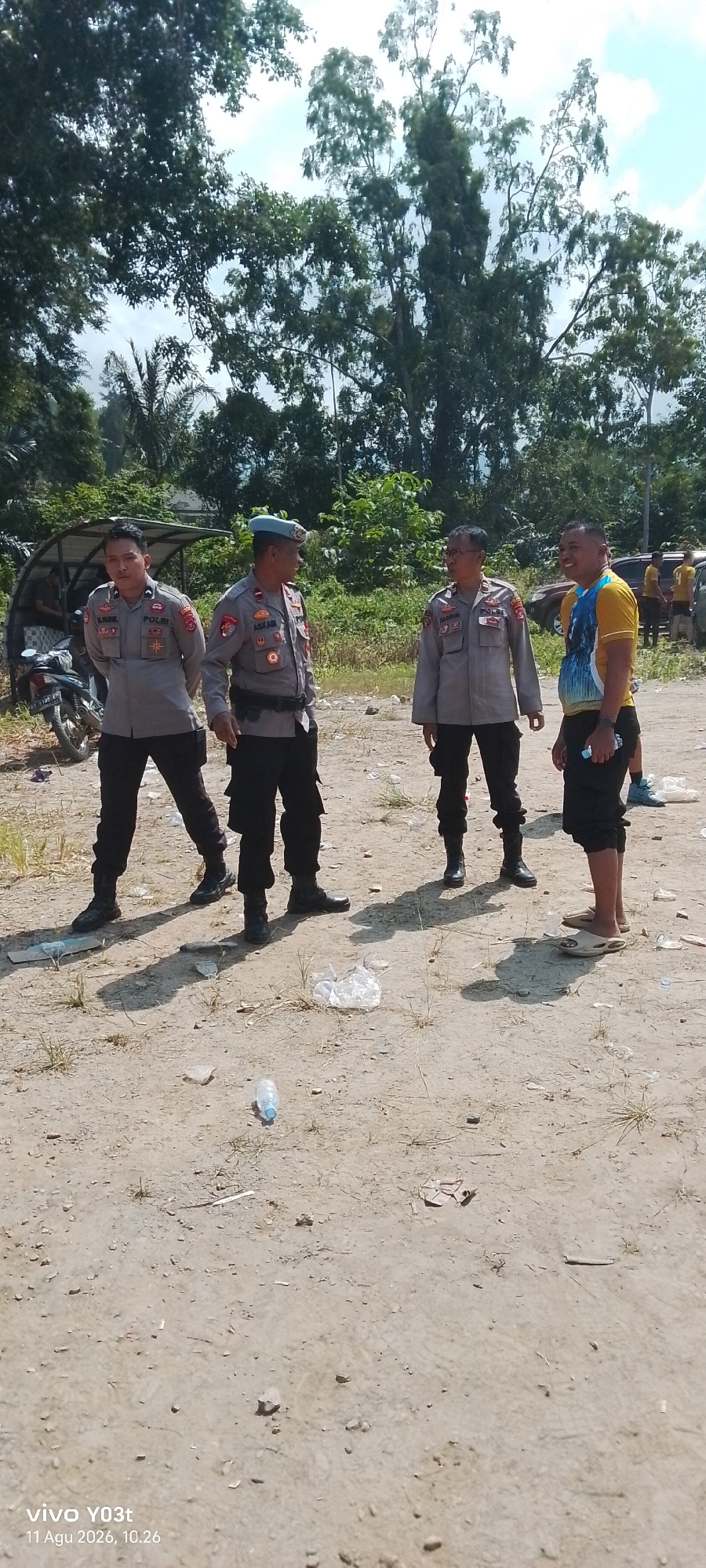 Four men are standing on a dirt ground outdoors. Three of the men are wearing police uniforms. One man is wearing a yellow and blue shirt. The man in the yellow and blue shirt is smiling and looking to his left. The man in the center of the group of uniformed men is wearing a blue beret and is looking to his right. The man to the left of the uniformed men is looking forward and down. The man to the right of the uniformed men is looking to his left. There are trees in the background and a motorcycle is parked on the left side of the image. There is trash on the ground. The date stamp on the image reads "11 Agu 2026, 10.26."