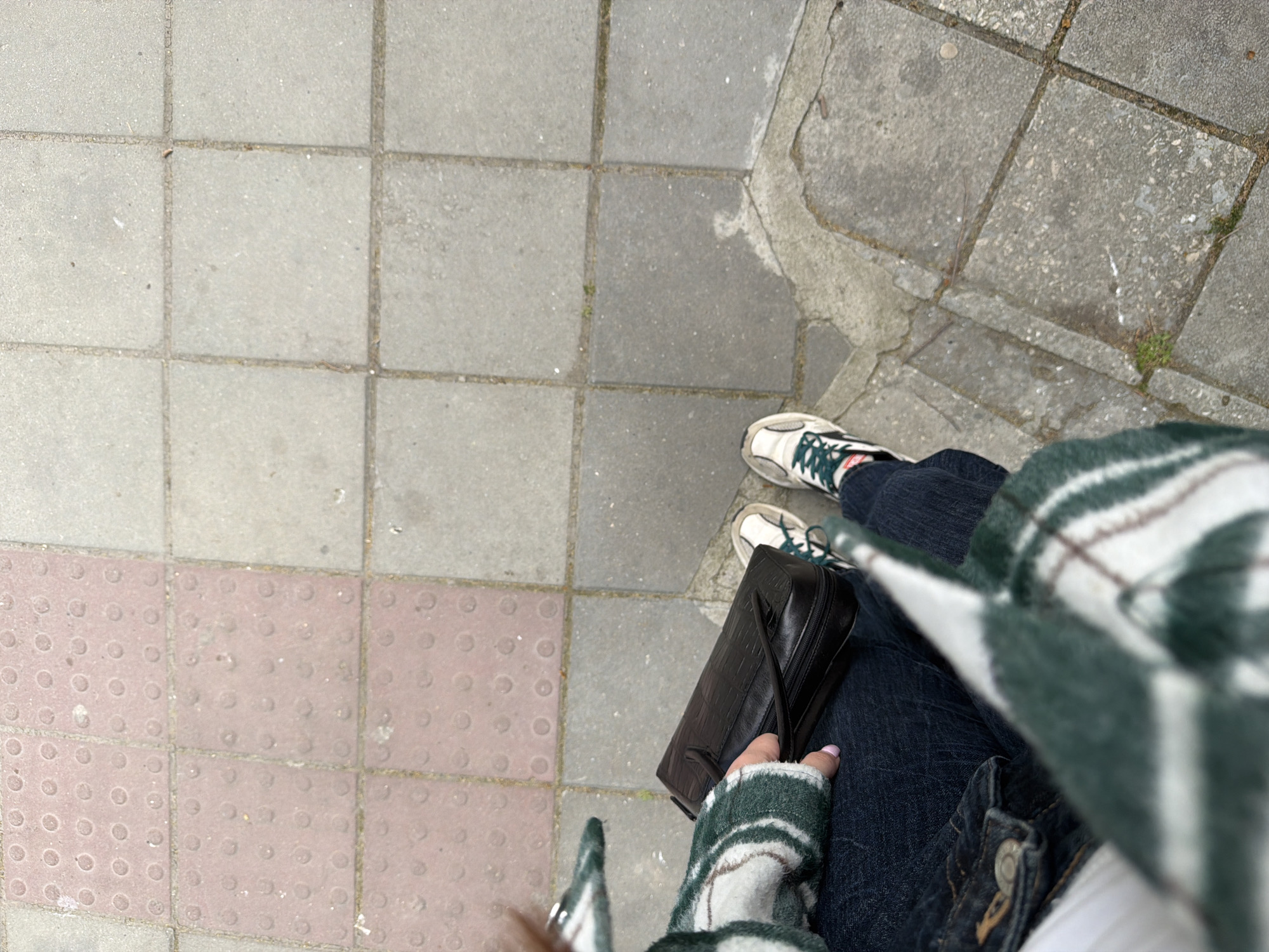 A downward shot shows a person standing on a sidewalk composed of grey and red square paving stones, likely in Varna, Bulgaria, given the unique style of paving. The person is wearing dark-wash jeans, white sneakers with green detailing, and a white and green plaid jacket. A black handbag rests on the person's leg. One hand is visible, holding the handbag. The sidewalk is adjacent to a different section of paving stones that are grey and uneven.