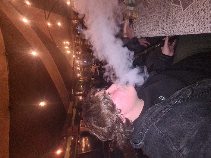 A person exhales a cloud of white smoke, likely from a hookah. The person, wearing a dark denim jacket, is lying sideways, facing right. The backdrop shows a long, dim restaurant or bar interior with tables and patrons visible in the distance. The lighting is soft and warm. A decorative ceiling with a curved design is also visible.