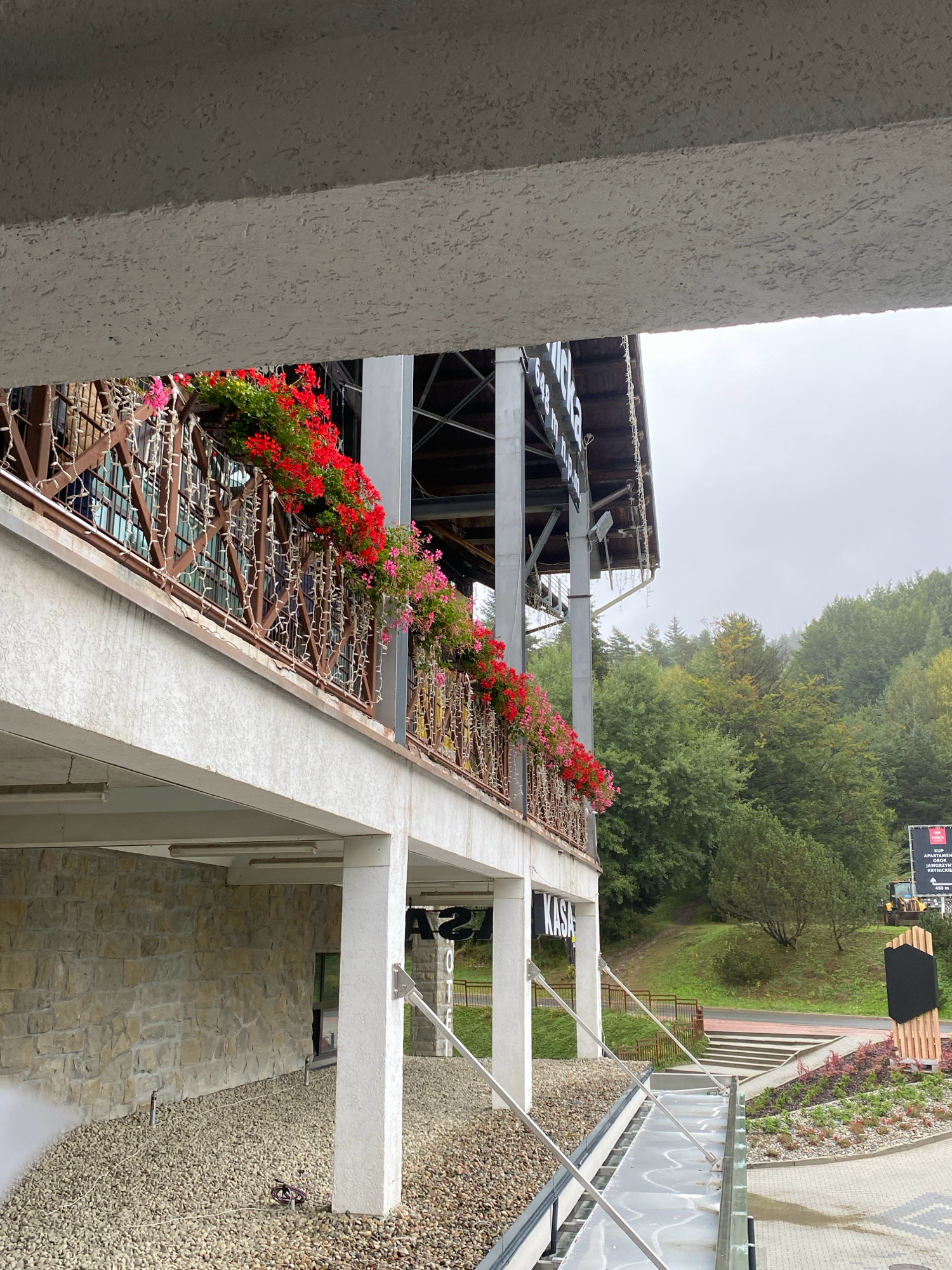 The image captures a view from an elevated perspective, looking towards a building with a balcony adorned with vibrant red and pink flowers. The building's facade features stone-like cladding on the lower level and white columns supporting an upper balcony. A sign reading "KASA" is visible on the building. Beyond the building, a lush green forest covers rolling hills under a cloudy sky. In the background, a yellow construction vehicle and a billboard are visible. The foreground shows a walkway made of glass or translucent material, supported by metal railings, leading away from the viewer. The overall scene appears to be in a natural or semi-rural setting, possibly a tourist area, given the signage and landscaping. It is located in Krynica-Zdrój, Poland.