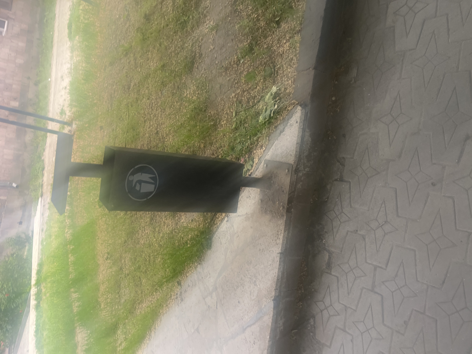 The image displays an outdoor scene, likely a public space or pathway, with a dark-colored, rectangular trash can as a prominent subject. The trash can is mounted on a concrete base and features a white circular symbol on its side, depicting a stylized person dropping an item into a bin, indicating its function as a waste receptacle.
Adjacent to the trash can is a concrete sidewalk characterized by a distinct repetitive geometric pattern, resembling star-like or interwoven shapes. To the left of the trash can and the sidewalk, there is a stretch of ground covered with sparse green grass and bare soil. In the background, a light-colored building, possibly constructed from stone or brick, is partially visible, along with hints of another path or unpaved area.
The scene is illuminated by natural light, suggesting it is daytime. The light appears somewhat diffused, possibly due to an overcast sky or haze. Some surfaces, including parts of the ground and pavement, show signs of dampness. No people or active events are captured in the frame; it is a static environmental shot. The setting appears to be a practical, utilitarian public area in Yeghegnadzor, Armenia.