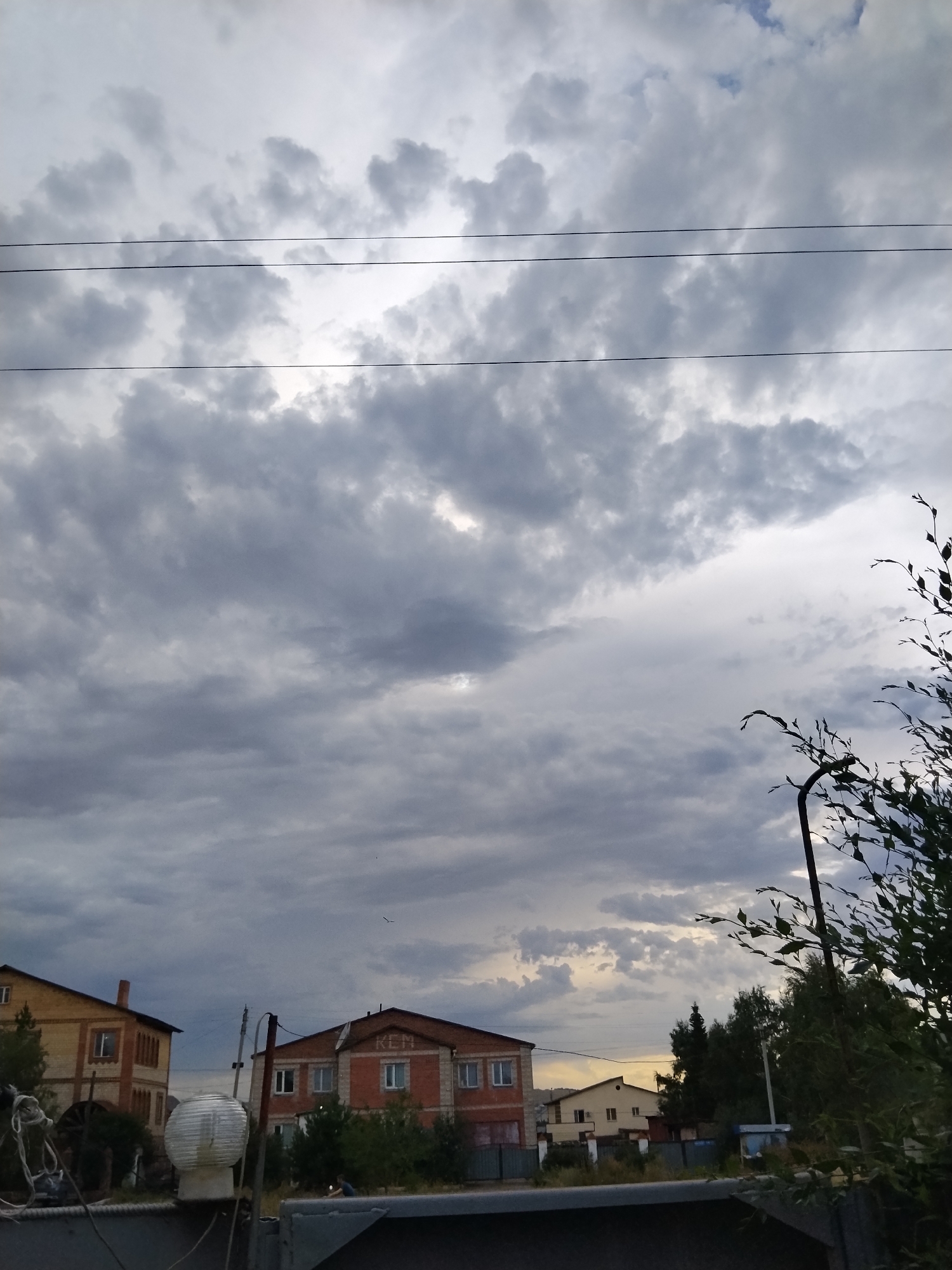 A low-angle, vertical shot captures a cloudy sky over a residential area in Kokshetau, Kazakhstan. Three power lines are strung across the upper portion of the frame, traversing a sky filled with dramatic, dark gray clouds interspersed with patches of lighter gray and white. The clouds appear heavy, suggesting an impending storm or overcast weather.
Below the sky, two brick houses are visible. The house on the left is a two-story structure with a light yellow brick facade and red brick accents around the windows and corners. It has a prominent chimney. The house on the right is also two stories, predominantly red brick with white-framed windows. The letters "KLM" are visible on its facade. In front of these houses, trees and other vegetation are sparsely scattered, and various objects, including what appears to be a round, white lamp or light fixture, are present in the foreground, partially obscuring the view of the houses. To the far right of the image, dark green foliage from a tree branches into the frame from the bottom. A faint silhouette of a bird can be seen in the sky above the houses. The overall impression is a somewhat moody, atmospheric scene of a residential neighborhood under a heavy sky.