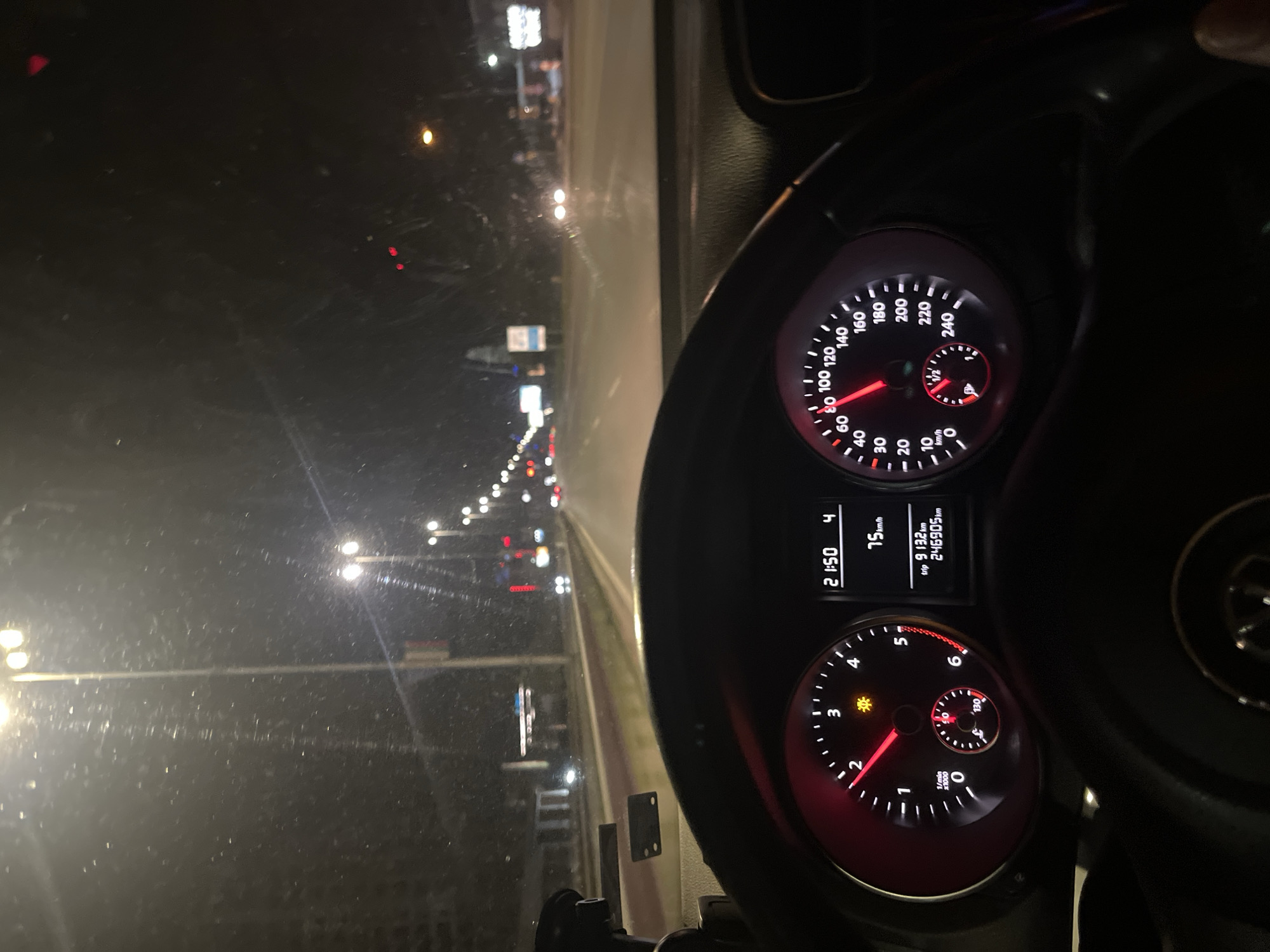 The image shows a view from inside a car, focusing on the dashboard and the road ahead at night. The car's speedometer indicates a speed of approximately 75 km/h. The tachometer shows the engine speed at roughly 2200 RPM. The odometer reads 246905 km. The dashboard clock displays the time as 21:50. The road ahead is illuminated by streetlights, and there are other vehicles visible. The setting is an urban environment with street lights and buildings visible in the periphery. Although the photograph provides no clues to the location, the prompt asks that the scene is located in Sofia, Bulgaria.