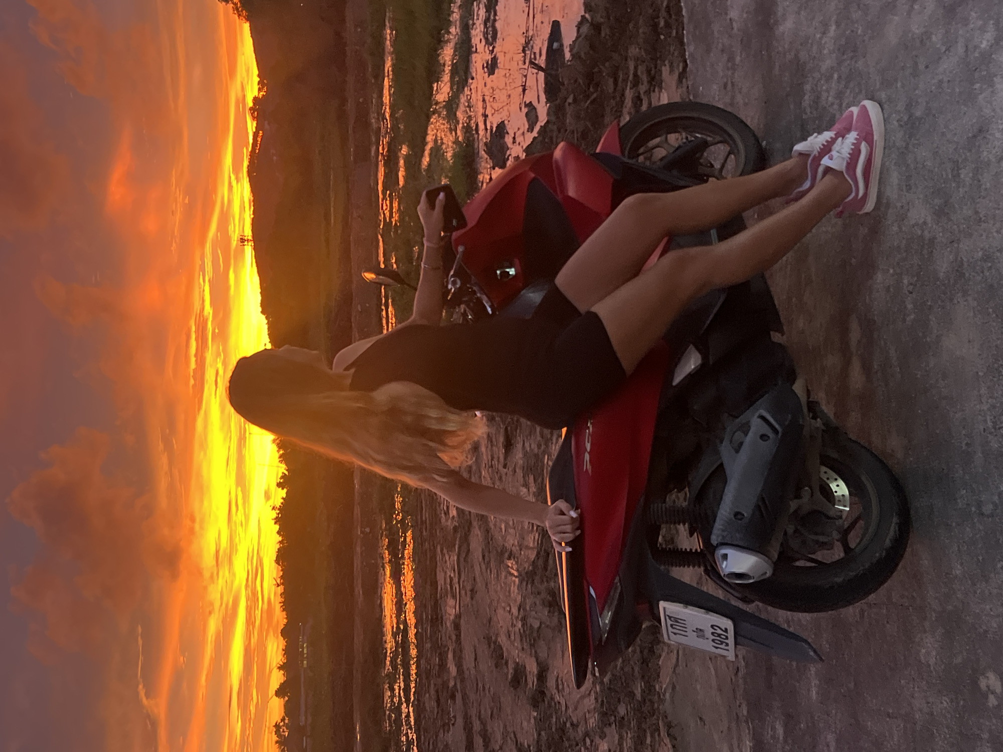 A young woman with long blonde hair sits on a red scooter, facing away from the camera, at sunset in Thep Krasatti, Thailand. The sky is ablaze with fiery orange and yellow clouds. The ground in front of the scooter is muddy and reflective, with patches of water. In the background, low hills are visible, covered in trees. The scooter has a license plate that reads "1กค 1982". The woman is wearing a black dress and pink Vans sneakers. The scene is set on an unpaved surface, possibly a dirt road or field.