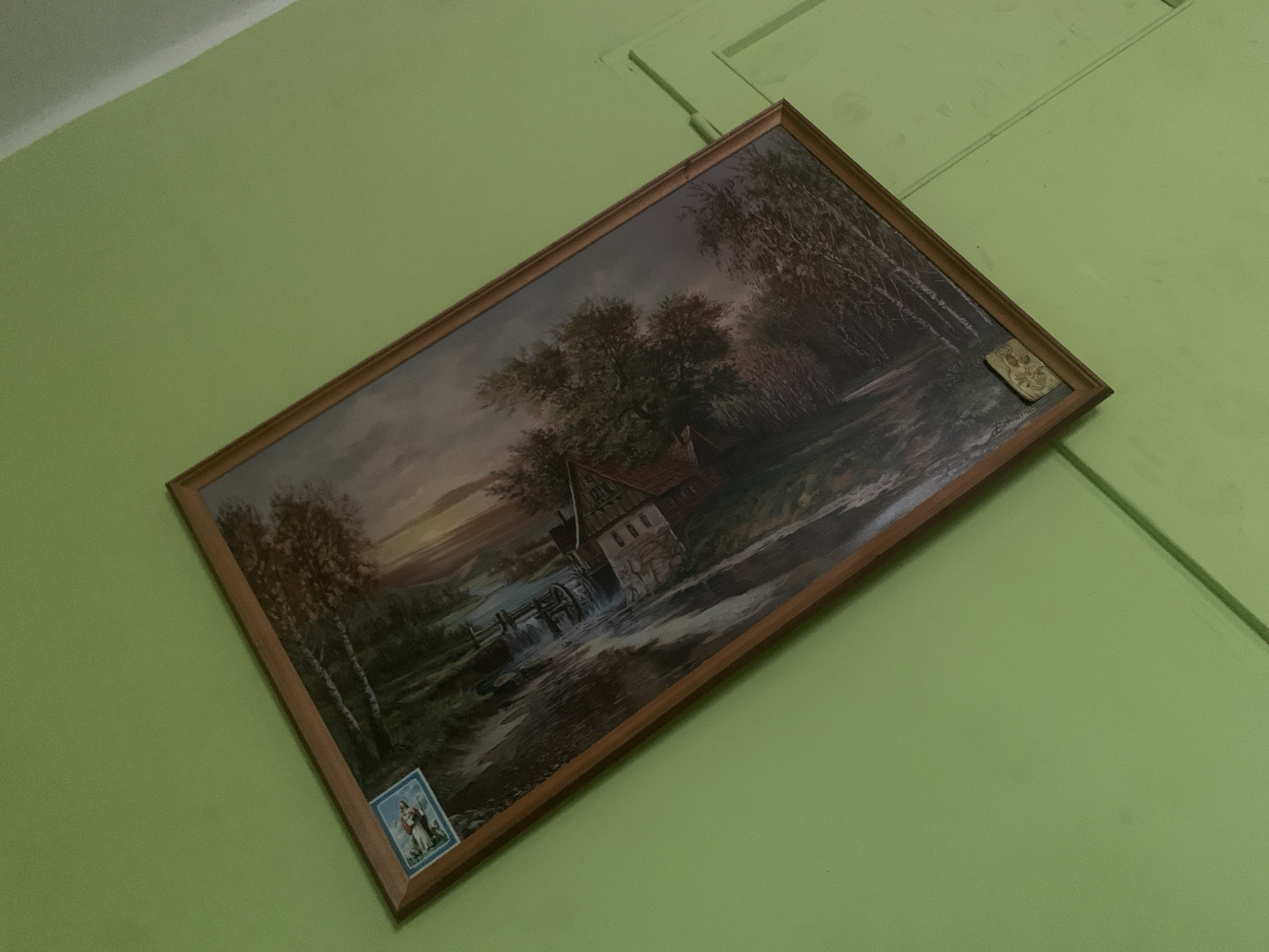 The image shows a framed painting hanging on a light green wall. The painting depicts a pastoral scene with a mill by a river, surrounded by trees. A small, rectangular blue and white religious image is affixed to the upper left corner of the painting's frame. A small, rectangular religious medal is affixed to the lower right corner of the painting's frame. The painting is oriented diagonally in the frame. The room appears to be a domestic interior, possibly a home in Skarydzew, Poland.