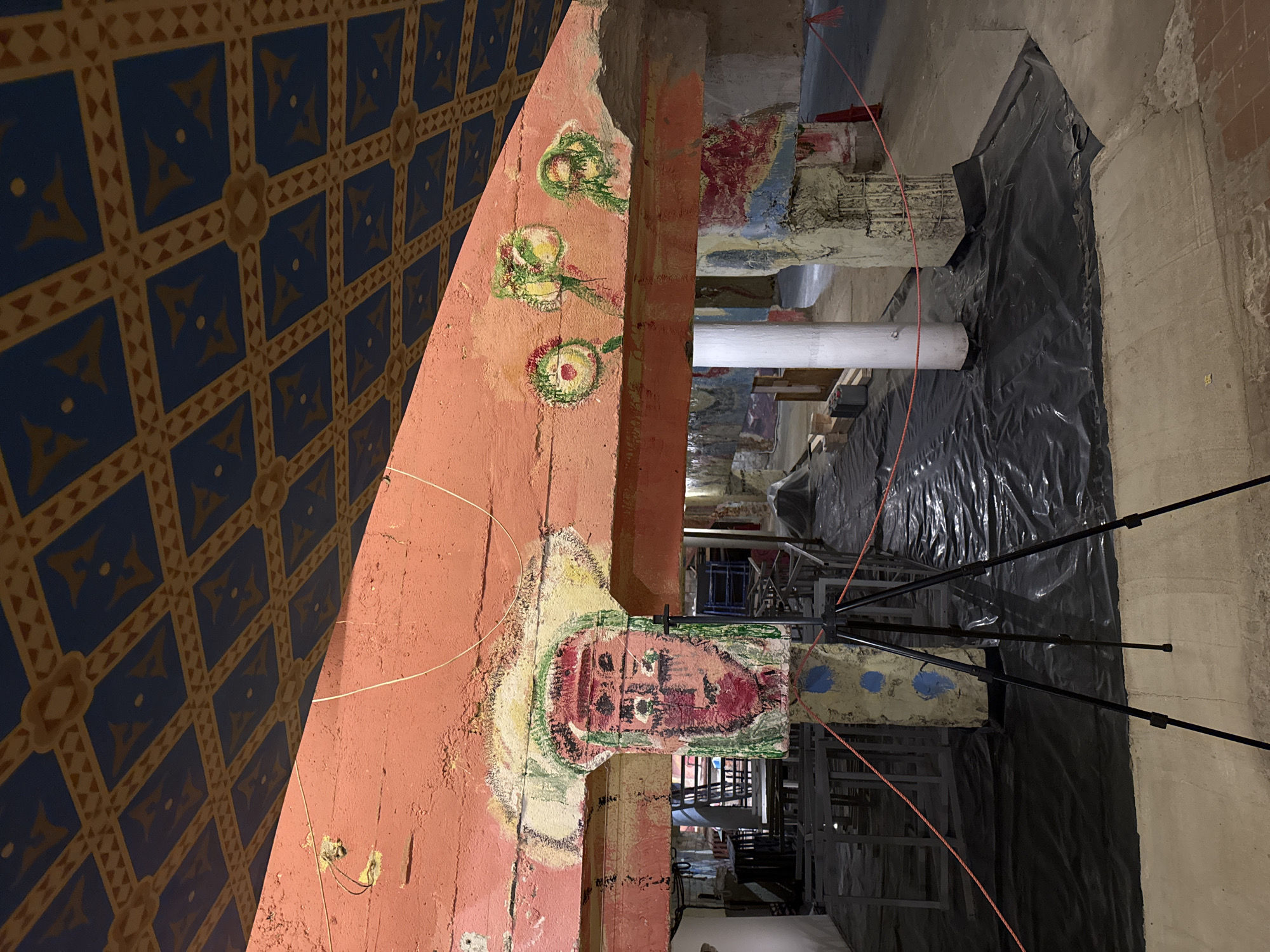 The image depicts an interior space, possibly a cellar or basement in Saint Petersburg, Russia, judging by the architectural details. A painted archway, decorated with geometric patterns in blue and gold on one side and crude floral and a possible facial image on the other, dominates the left foreground. The archway is painted in red and appears to be part of an old building's structure.
In the background, the space opens into a larger area, partially obscured by black plastic sheeting. Scaffolding and metal structures are visible, indicating ongoing construction or renovation. A white pipe runs vertically through the space.
A black tripod is placed in the center, suggesting photographic or surveying activity. The floor appears unfinished, and the overall impression is one of an area undergoing preservation or restoration. The crude artwork on the archway is notable, possibly indicating the location has historical significance.