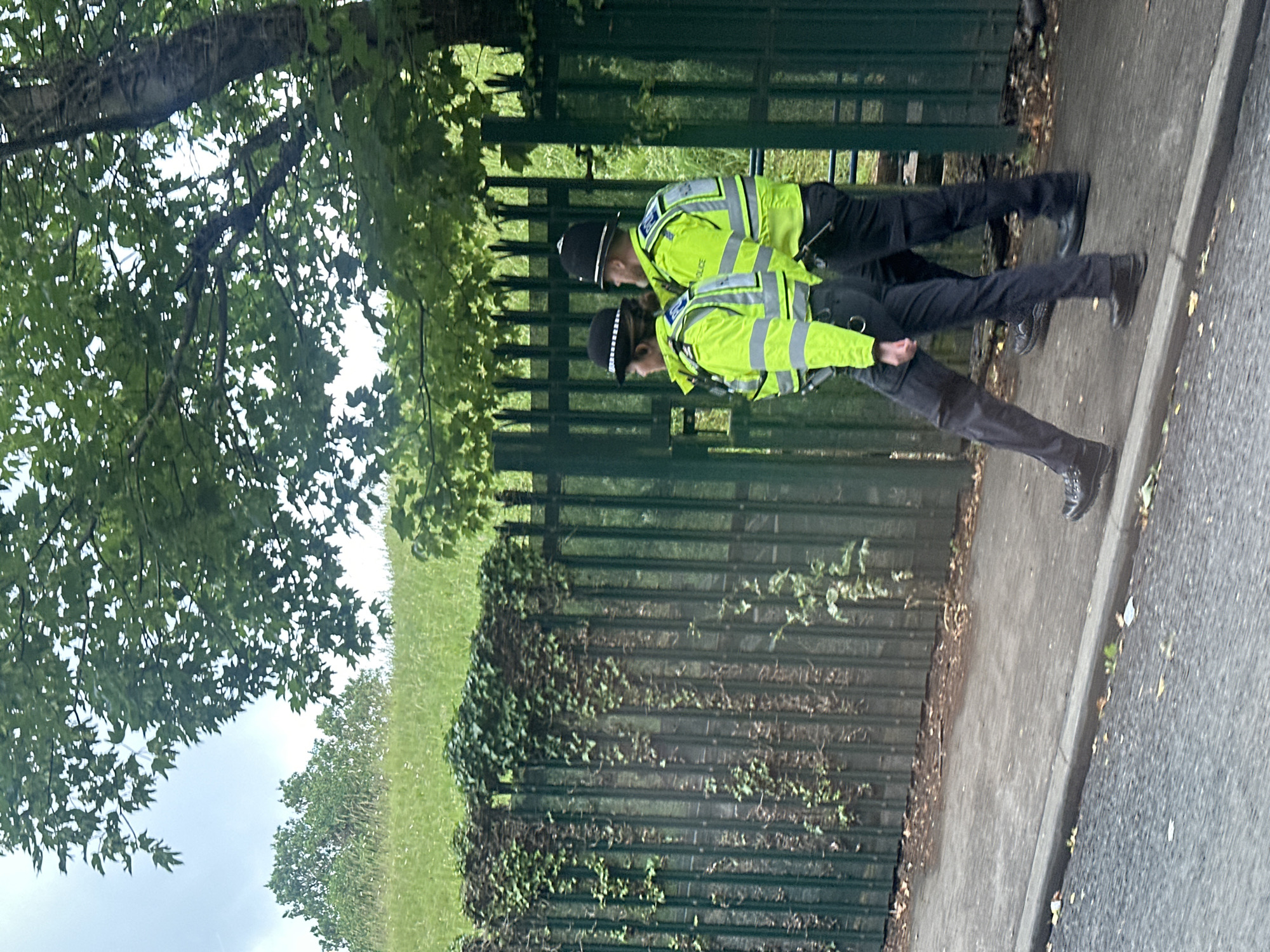 The image displays two uniformed British police officers, a male and a female, walking from left to right on a paved footpath in Epsom and Ewell, United Kingdom. The female officer, positioned to the left, wears a dark, brimmed police hat, a bright yellow high-visibility jacket with reflective silver stripes, dark trousers, and black boots. The male officer, positioned to the right and slightly behind, wears a traditional black British police custodian helmet, a matching bright yellow high-visibility jacket with "POLICE" visible on the back, dark trousers, and black boots, with a black equipment holder attached to his belt. Their action is a coordinated walk along the path. Immediately to their right, running parallel to the footpath, is a tall, dark green metal fence, partially obscured and heavily overgrown with green ivy and other foliage. Beyond the fence, a gentle green grassy slope ascends, topped by several trees under a bright, overcast sky. A section of a dark grey asphalt road is visible in the immediate foreground.