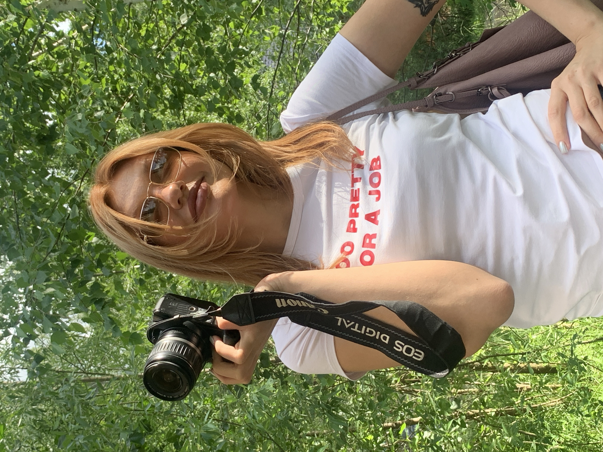 A young woman with shoulder-length blonde hair and sunglasses holds a black Canon EOS digital camera in her left hand. She is wearing a white t-shirt with red text that reads "DO PRETTY OR A JOB". A brown shoulder bag is slung over her right shoulder. The woman is standing outdoors, with a background of lush green foliage. The scene appears to be a park or wooded area, possibly in the city of Chaikovsky, Russia, given the context provided.
