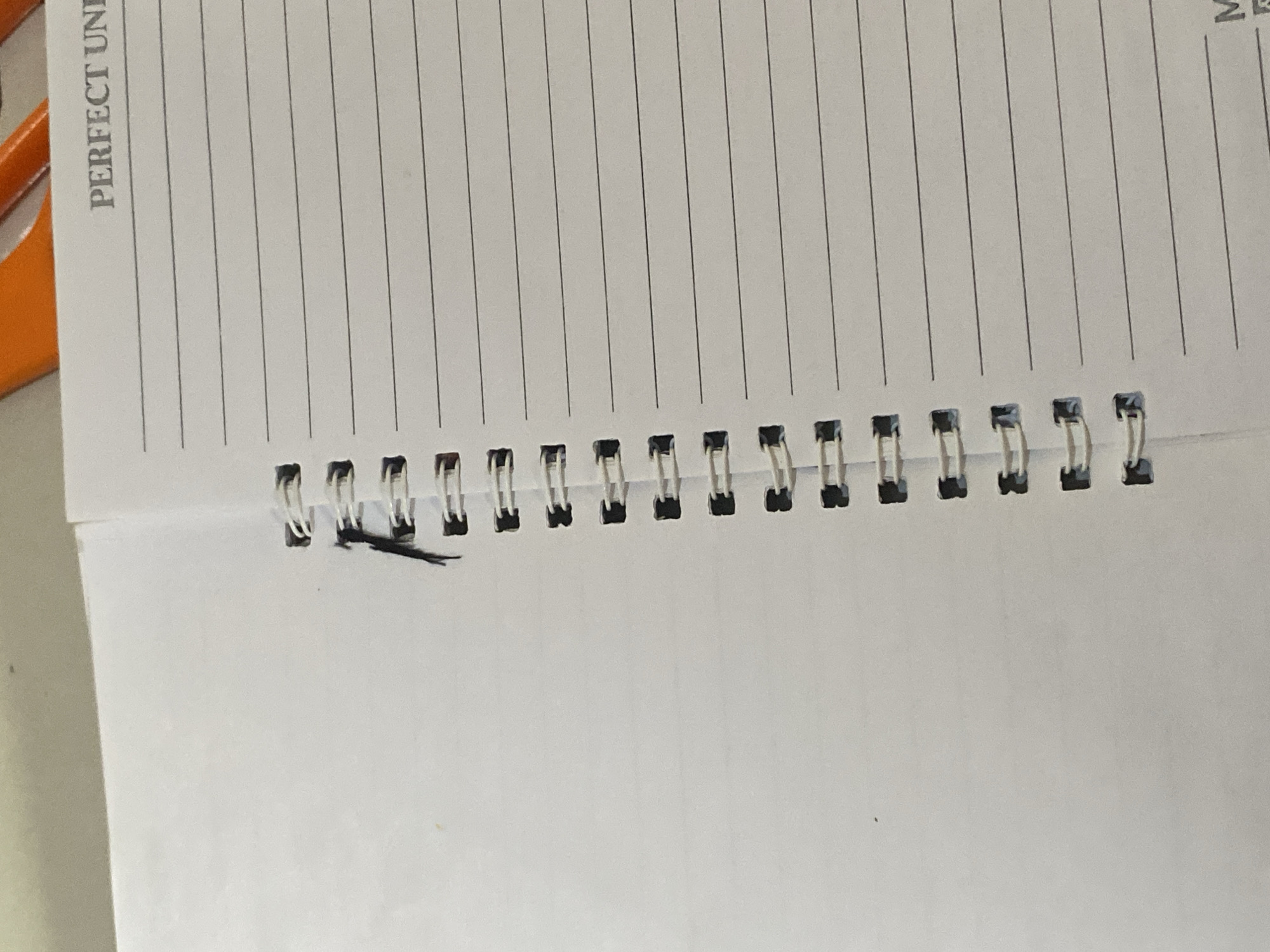 The image shows a close-up of an open notebook. The notebook has white lined pages and a silver spiral binding along the left edge. A small piece of black material is caught in the spiral binding. The text "PERFECT UNI" is visible in the upper right corner of the right page, suggesting the notebook is branded. The background is blurred and out of focus, with hints of an orange object at the top. The scene is a still-life, implying a stationary subject. There are no people, actions, or interactions depicted. The environment is a flat surface, likely a table. There is no information within the image to suggest a specific location in Jauro Yinu, Nigeria.