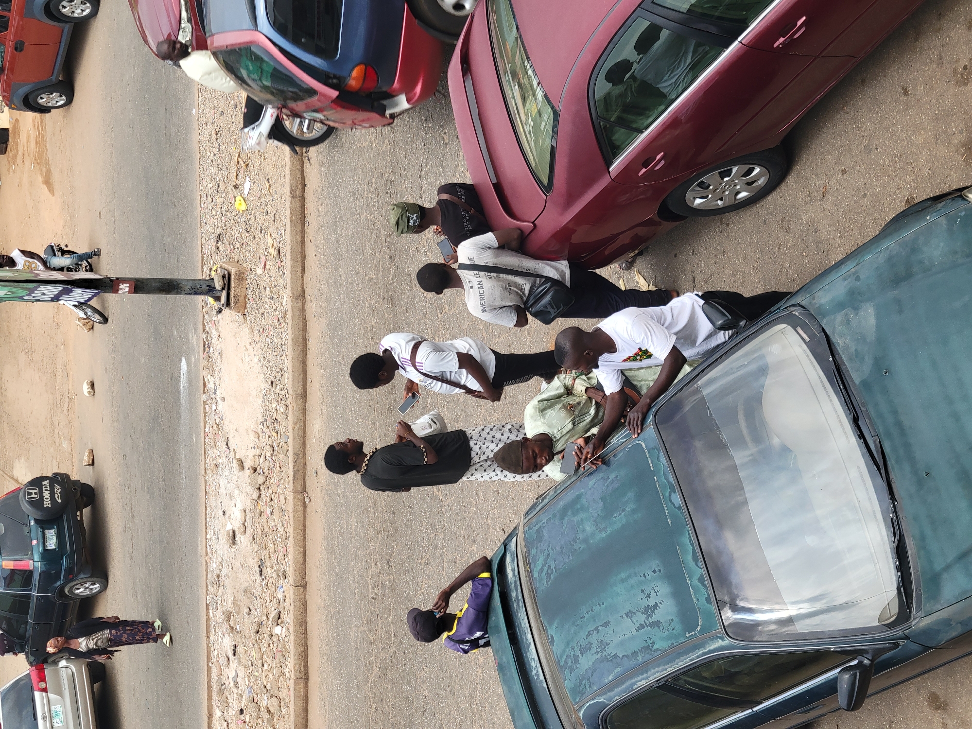 The image depicts a street scene in Jos, Nigeria, featuring a line of parked cars and a group of people gathered beside them. Six individuals are positioned near the cars, some of whom are holding or looking at mobile phones. The vehicles include a maroon sedan, a blue compact car, a dark green car, and a dark green Honda SUV. Other vehicles are visible in the background. The street is paved, with a dirt and gravel shoulder on one side. A street sign is visible on the side of the street.