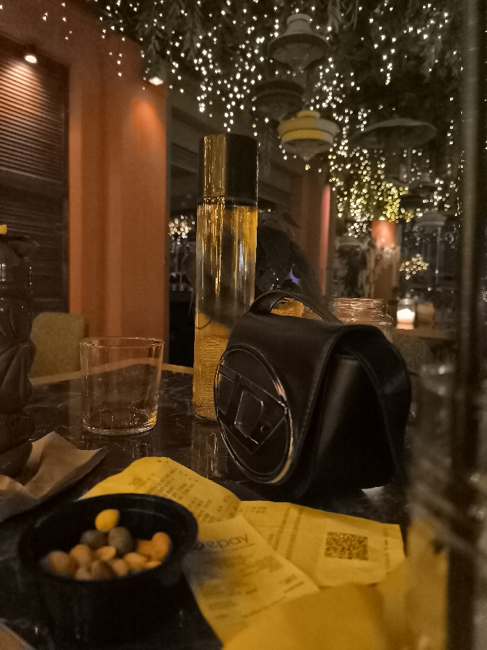 The image depicts an intimate, dimly lit indoor scene, likely a cafe, bar, or restaurant, taken from a low angle across a polished, dark table. The setting is characterized by warm, artificial light, primarily from numerous small, twinkling string lights suspended overhead, possibly intertwined with greenery, and diffuse orange lighting from walls and distant fixtures.
The main subjects on the table include a tall, clear bottle filled with a golden-yellow liquid, an empty clear glass tumbler, and a small black purse or bag with a prominent silver 'D' shaped logo, suggesting the brand Diesel. Scattered across the table are several yellow paper receipts, one visibly marked "epay" and another displaying a QR code. A small black bowl on the left contains various colorful, coated snacks or candies. Part of a dark, patterned object, possibly a decorative mug, is also seen on the far left.
In the soft-focus background, orange-colored structural elements and other hanging lamps are visible, contributing to the cozy atmosphere. The activity appears to be a casual evening gathering, with food and drink items suggesting a social occasion. The time of day is clearly night or late evening due to the reliance on artificial lighting. The location is Palaio Faliro, Greece, implying a local establishment. The overall mood is relaxed and inviting, with no people directly visible in the frame.