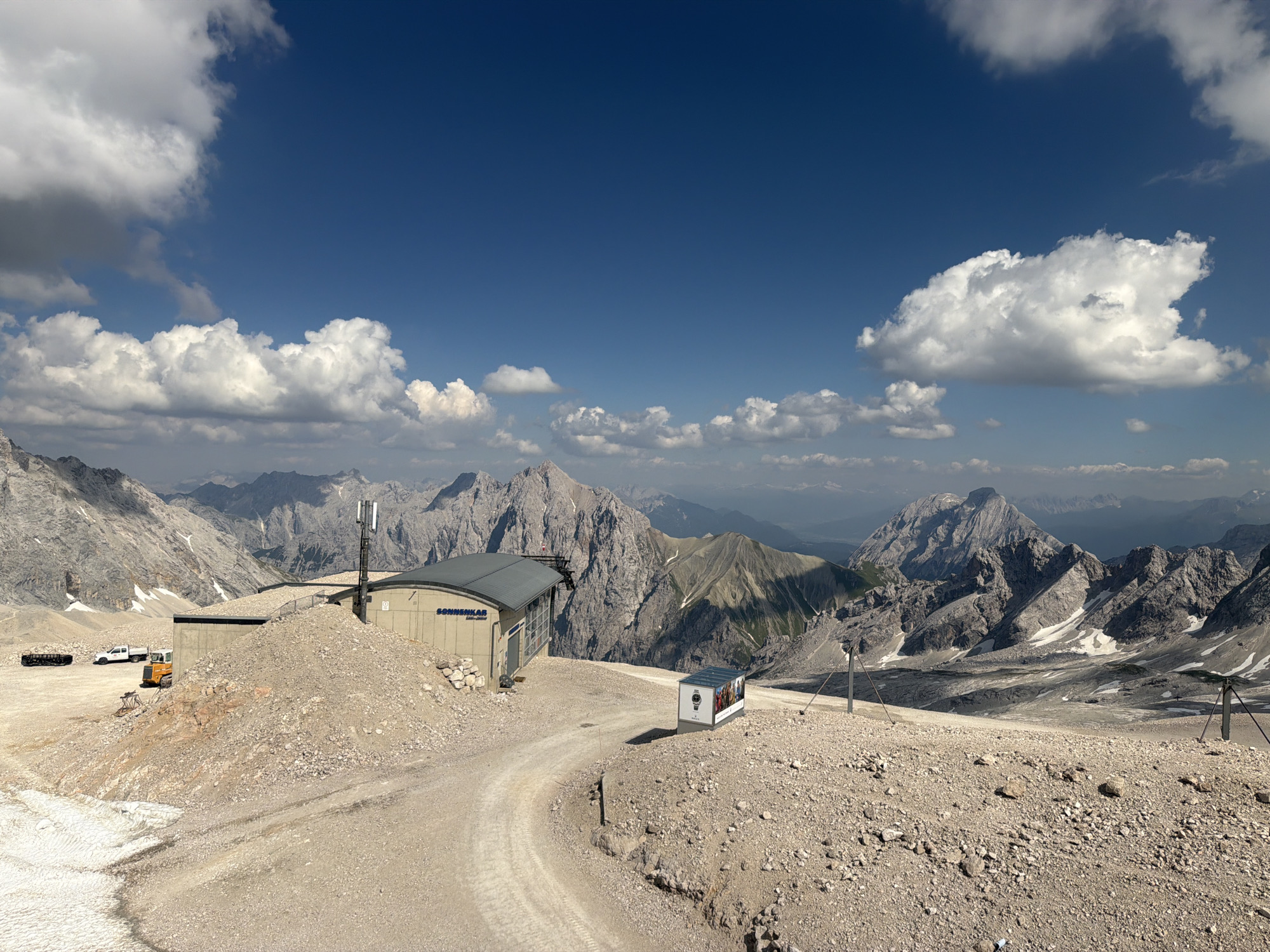 The image depicts a high-altitude mountainous landscape on a sunny day with scattered clouds. The foreground features a gravelly, rocky terrain with patches of snow. A winding dirt road leads towards a modern building with a gray roof, labeled "SONNENKAR" and "EISENBAHN" (railway). A telecommunications tower stands next to the building.
In the mid-ground, several vehicles are visible: a yellow bulldozer, a white pickup truck, and a track loader. The background is dominated by rugged, imposing mountain ranges with rocky peaks and some snow-covered slopes. The sky is a deep blue with fluffy white clouds, suggesting pleasant weather conditions, likely midday given the brightness. The overall scene conveys a sense of remote, natural beauty, with a touch of human infrastructure providing access or services. A small, refrigerated display box with advertisements is also present near the road.
The location is Garmisch-Partenkirchen, Germany, which is known for its alpine scenery and mountain railways. The presence of the "EISENBAHN" sign on the building further supports this context. There is no clear activity taking place, but the presence of vehicles suggests ongoing work or maintenance. The bright sunlight and clear skies evoke a feeling of openness and grandeur.