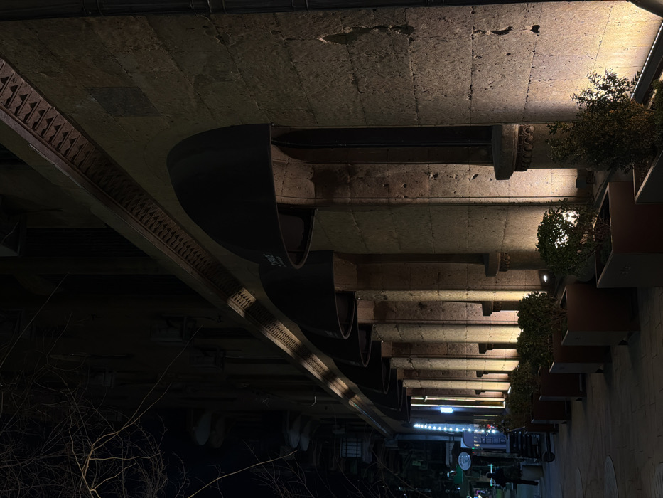 The image depicts the illuminated nighttime facade of a commercial building on Abovyan Street, Yerevan, Armenia. The structure is constructed of light-colored, rough-hewn stone, likely tuff, exhibiting visible mortar lines and some surface imperfections. Six arched openings are visible along the ground floor, each fitted with a dark, semi-circular fabric awning; the leftmost visible awning bears the white text "JAZZVE." A decorative frieze with a repeating geometric pattern runs horizontally above the awnings. Vertical, warm-toned spotlights positioned at ground level illuminate the stone surfaces between each archway, creating distinct shafts of light and shadow. In front of the building, a row of approximately six dark, rectangular planter boxes contains small, green shrubs. The sidewalk in the foreground is visible. In the distant left background, a lone, indistinct human figure is partially visible on the street, near other illuminated establishments and bare tree branches. The scene is static, with no overt actions or interactions.