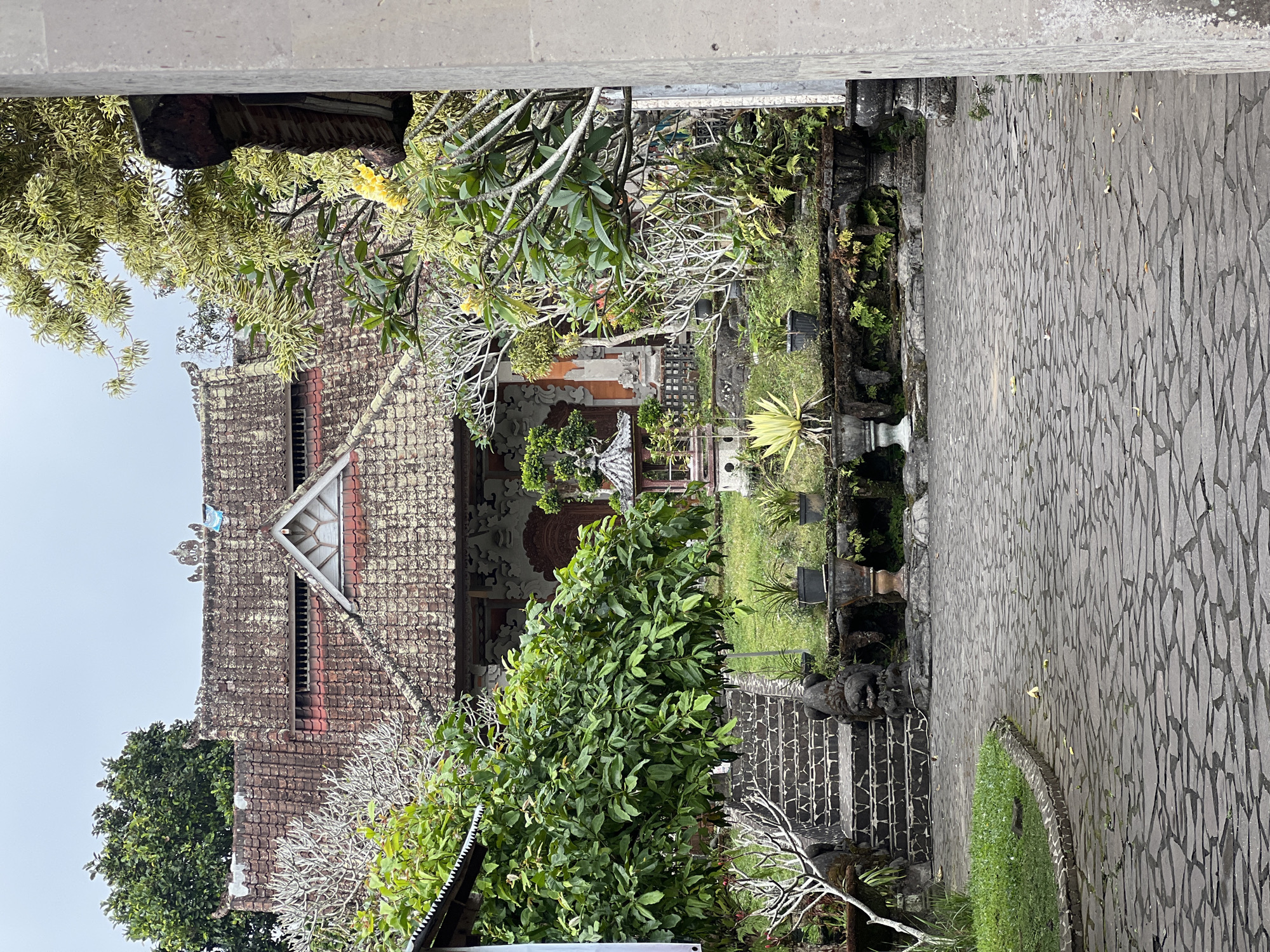 The image shows the exterior of a traditional building with a tiled roof in Rendang, Indonesia. The building is surrounded by lush greenery, including trees and potted plants. A stone pathway leads towards the entrance, with a set of stone steps to the left. The architecture appears to be of Balinese influence, with ornate carvings visible on the building's facade and decorative elements in the garden. The sky is overcast, suggesting a potentially humid or rainy day. There are no people visible in the scene.