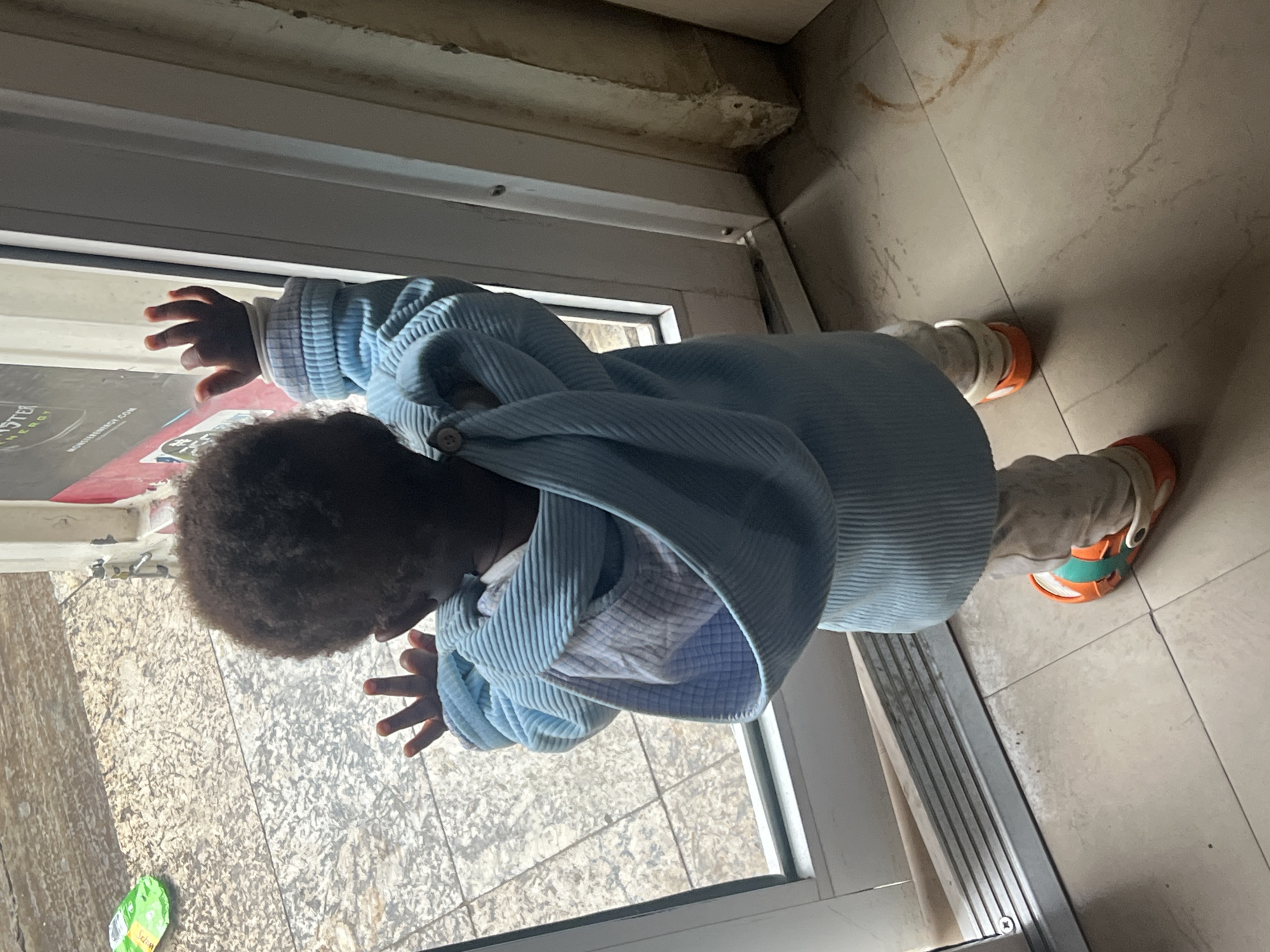 A child stands near an open doorway, with one hand touching the glass and the other outstretched. The child wears a light blue jacket, light pants, and orange Crocs. The doorway leads to a tiled floor. A package of candy or gum is visible on the ground outside the door. The location is Gura Top, Nigeria.