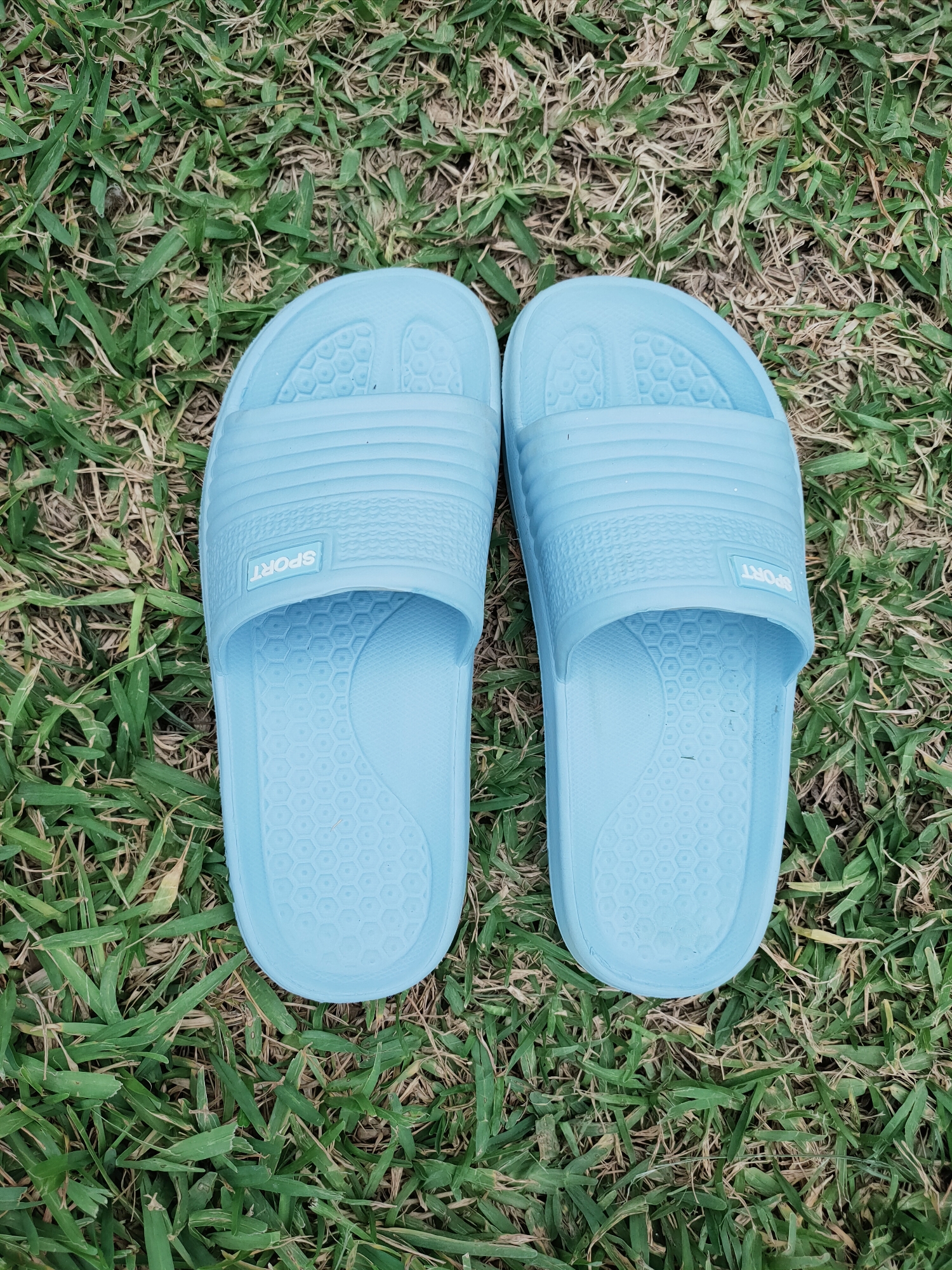 Two light blue rubber slides with the word "SPORT" imprinted on them are positioned side-by-side on a patch of dry grass. The slides are oriented with the toe pointing upwards, and the interior of each slide features a hexagonal patterned sole. The grass is predominantly brown with some patches of green, suggesting it is either dry or a mixed lawn. The lighting indicates an outdoor setting, likely during the day. No people or other objects are visible in the frame. The specific location in Mrezga, Tunisia cannot be determined from the image alone.