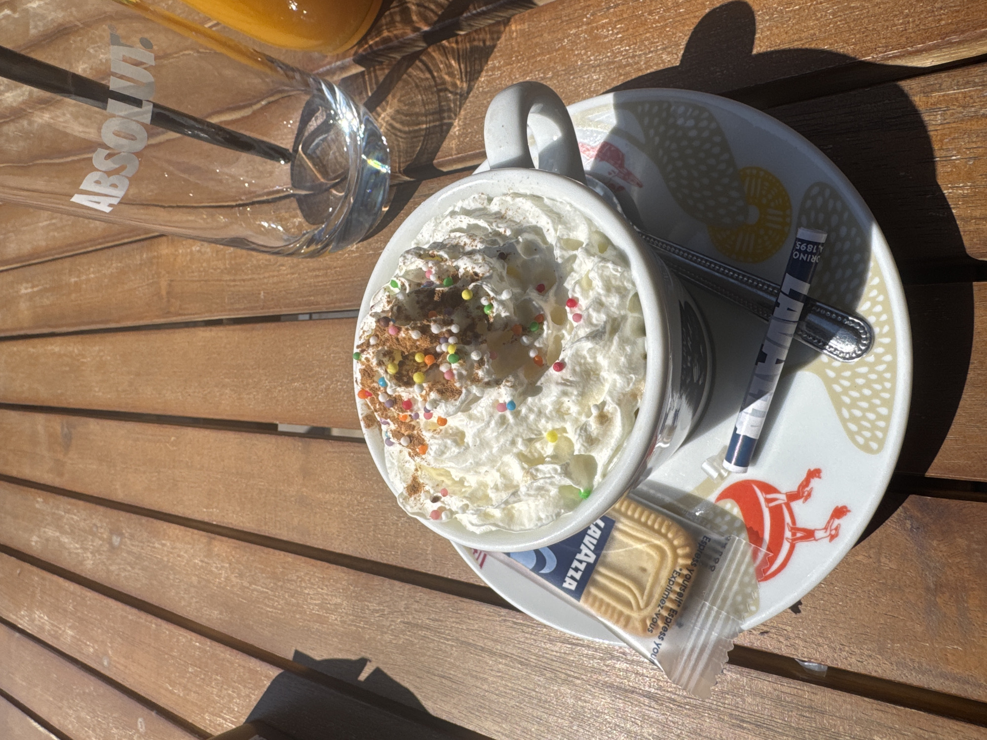 A cup of coffee topped with whipped cream and sprinkles sits on a saucer next to a glass of orange juice on a wooden table. A packet of Lavazza biscuit, a sugar packet, and a spoon are on the saucer as well. The glass has "ABSOLUT." printed on it. The picture was taken on an outdoor table, possibly in Byala Slatina, Bulgaria.