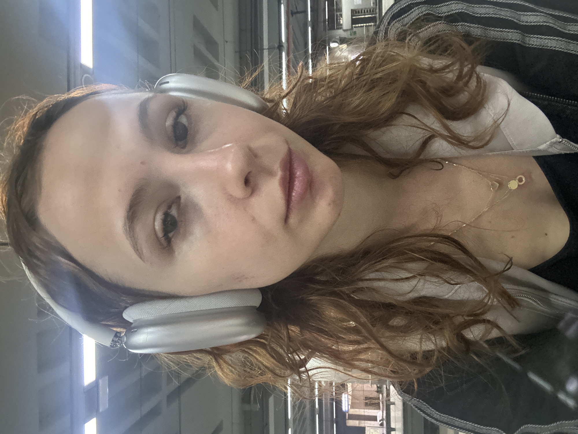 A woman is shown in a close-up selfie-style photograph. She is wearing silver over-ear headphones and a light-colored top with a gold necklace. The background appears to be an indoor public space with metallic beams and windows, suggesting a station or transport hub. There are no other people visible. No specific landmarks identifying Belgrade, Serbia are visible in the frame. The lighting is bright and somewhat harsh.