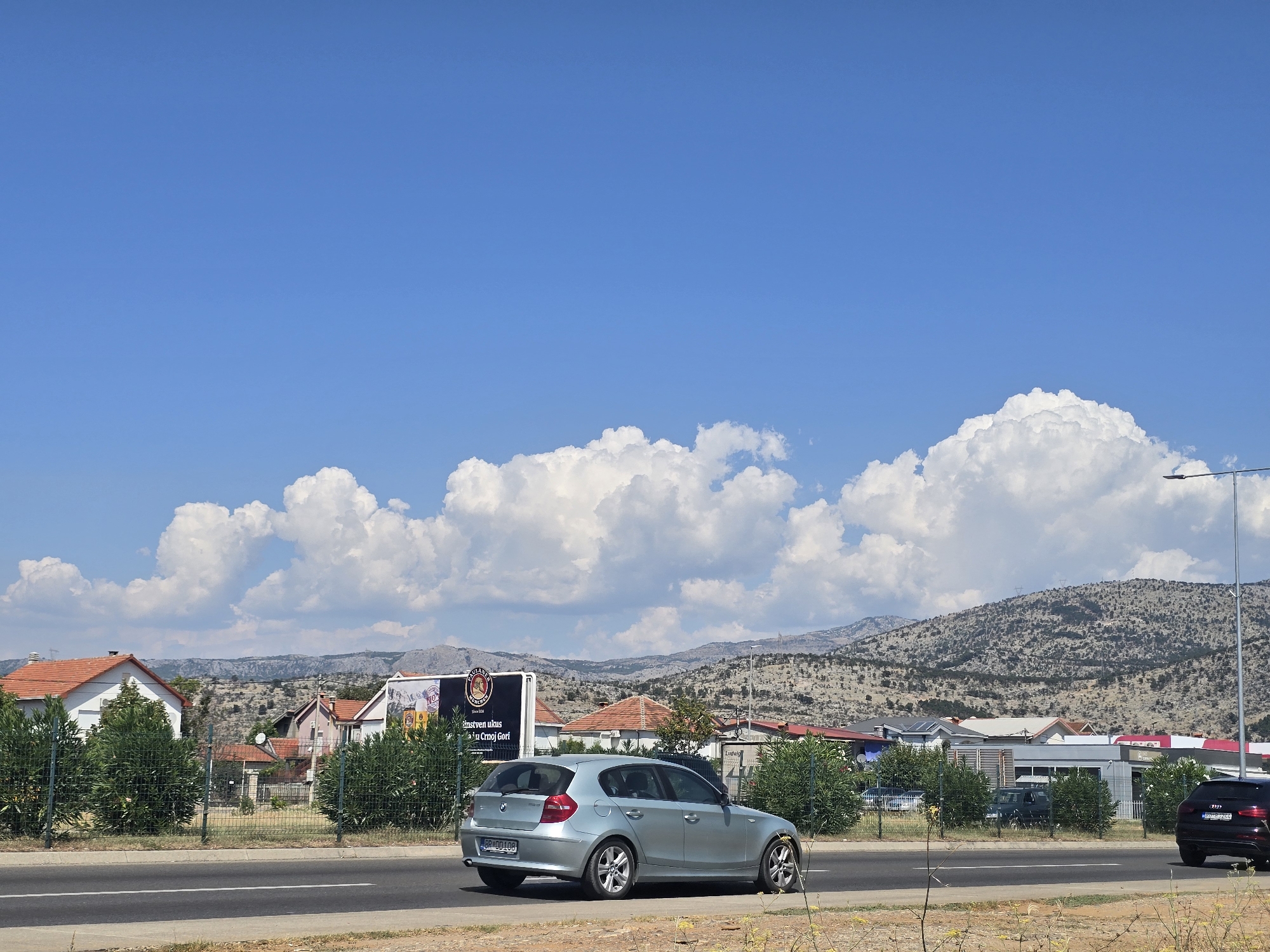 The image captures a sunny day with a bright blue sky dotted with fluffy white clouds. A light blue hatchback car is driving on a paved road in the foreground, with its rear facing the camera. In the background, there are several houses with red-tiled roofs, some commercial buildings, and a billboard. The landscape behind the buildings consists of dry, rocky hills. A street lamp is visible on the right side of the image. The overall scene suggests a suburban or exurban area on a clear day.