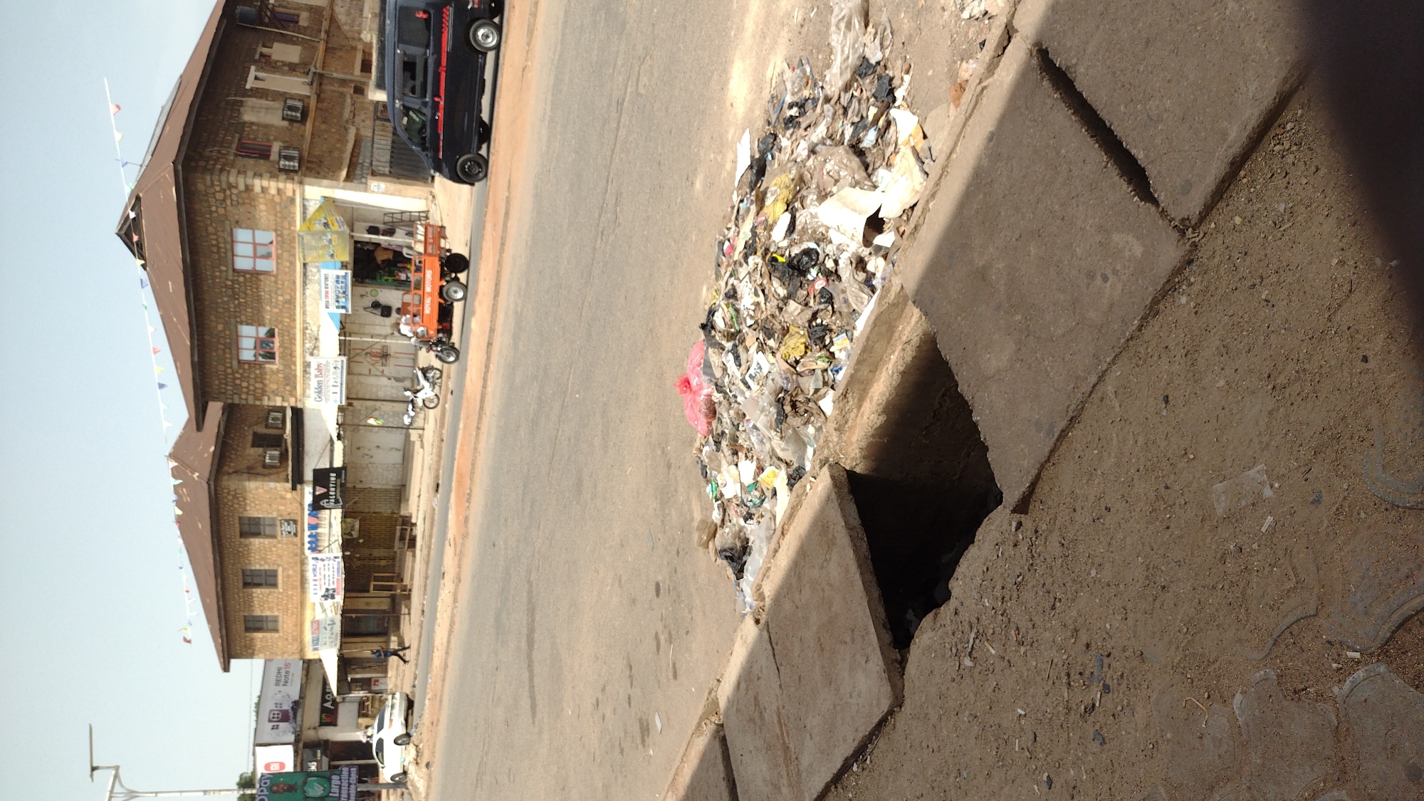 The image shows a street scene in Jos, Nigeria. A storm drain is filled with trash and debris. Buildings and vehicles line the opposite side of the street.