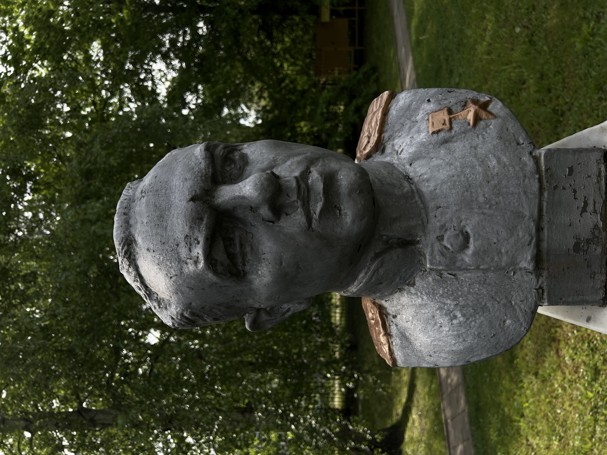 A bust statue stands outdoors in Goryachiy Klyuch, Russia. The statue is grey, roughly textured, and depicts a man with short hair wearing a military uniform. The uniform has gold-colored epaulettes and a gold-colored star and rectangular symbol on the left chest. The statue is positioned against a backdrop of green trees and grass. A concrete pathway is partially visible in the background.