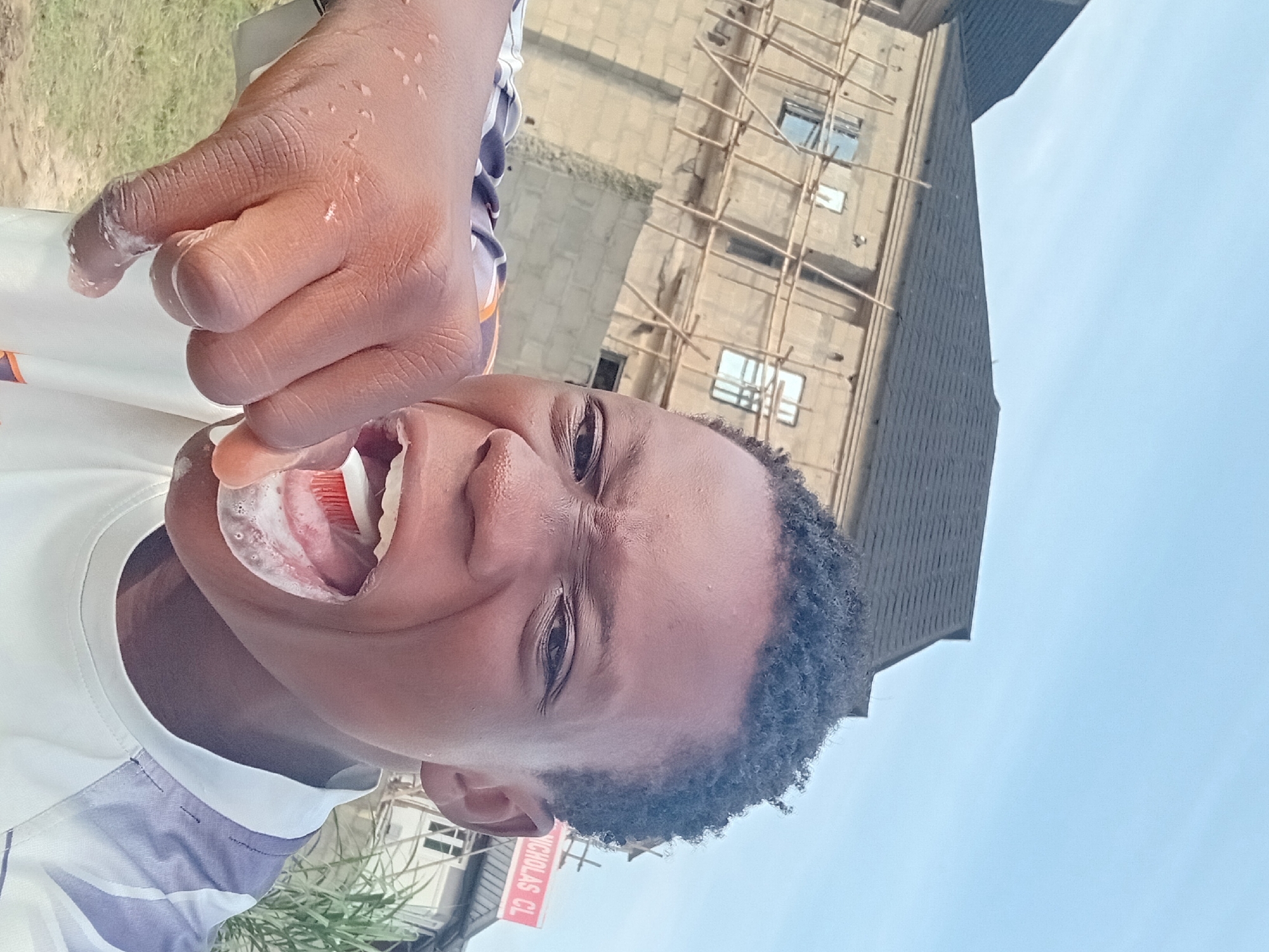 A close-up, selfie-style shot captures a young person brushing their teeth outdoors. The person's mouth is open, revealing a toothbrush with red bristles and white foam, actively being used. Their hand, with visible water droplets and foam, holds the toothbrush. The background features a building under construction, with scaffolding and exposed brickwork, suggesting an urban or suburban setting. The sky is a pale blue, and some green foliage is visible on the right side. A red and white sign with partially visible text "NICHOLAS CL" is mounted on the building. The overall lighting suggests daytime.