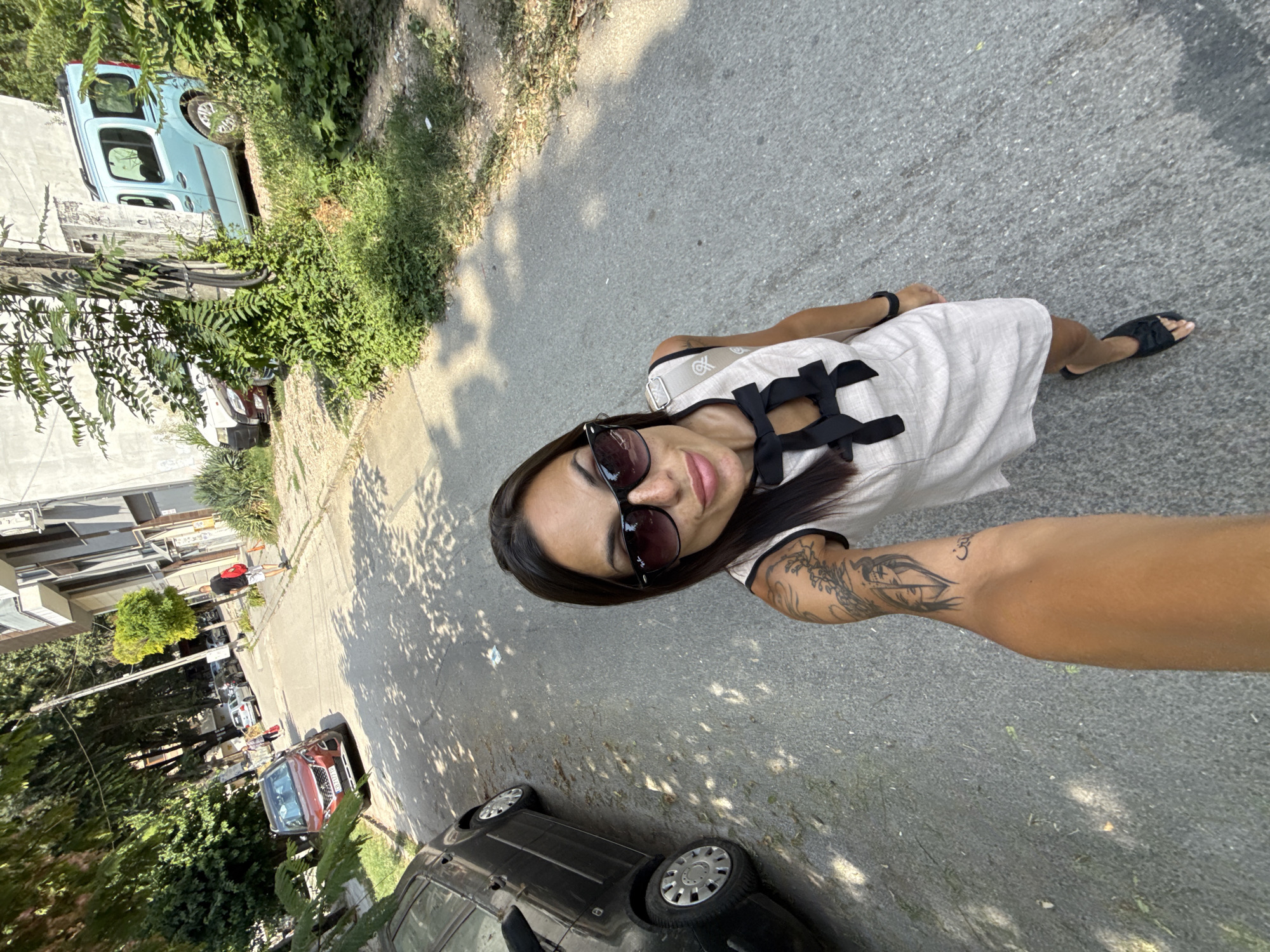 A woman in a light-colored dress with black bows, sunglasses, and black sandals walks down a street in Pleven, Bulgaria. She is taking a selfie, with her arm and a tattoo on her arm visible. A black car is parked on the left side of the street, and a red SUV is parked further up. The street is paved and lined with trees and buildings. A light blue van is parked on the right side of the street behind some greenery. A man in a black t-shirt and white shorts is walking away from the camera on the sidewalk. The scene appears to be a sunny day.