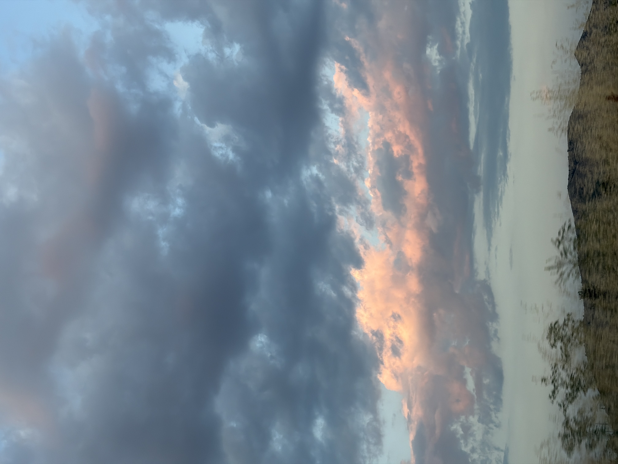 The image captures a landscape scene with a dramatic sky. Dark, textured clouds dominate the upper portion of the frame, interspersed with lighter, wispy formations. A band of pink and orange clouds runs horizontally across the mid-ground, suggesting a sunset or sunrise. Below the clouds, a blurry, out-of-focus foreground shows the tops of vegetation and the silhouette of rolling hills. The overall impression is one of natural beauty and atmospheric conditions.