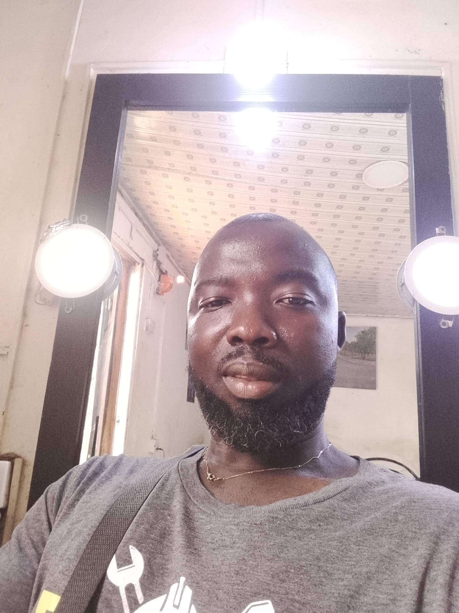 A head-and-shoulders selfie shows a dark-skinned man with a closely cropped, slightly graying beard and a gold chain around his neck. He wears a gray t-shirt with a graphic design featuring a wrench. The man appears sweaty. Behind him is a mirror framed in black, and two round lights are mounted on either side of the mirror. The reflection in the mirror reveals a patterned ceiling and a window to the left, allowing a view of a light-colored wall. A small framed picture is visible in the reflection on the right. The location is in Gwong, Nigeria.
