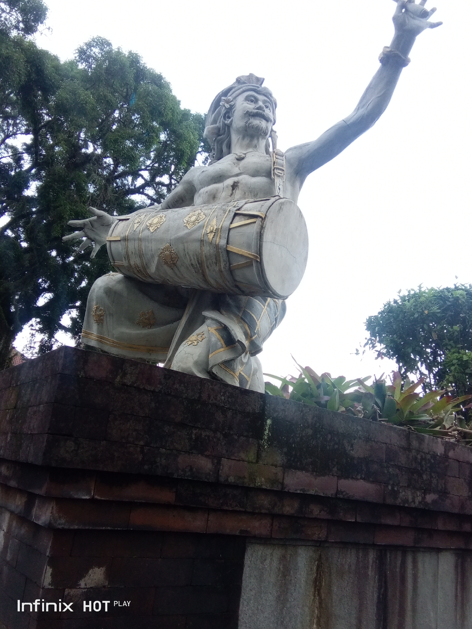A low-angle outdoor photograph shows a stone statue of a man playing a drum. The statue is of a man with a mustache and long hair, wearing a headdress. He is seated and holding a large drum horizontally across his lap, with his hands positioned as if playing it. The drum is decorated with gold accents. The statue is weathered and appears to be old. Behind the statue, large green trees are visible against a bright, overcast sky. In the foreground, a brick wall forms the base of the statue's pedestal. The scene appears to be a public space, possibly a park or temple. The bottom left corner of the image displays the text "Infinix HOT PLAY".