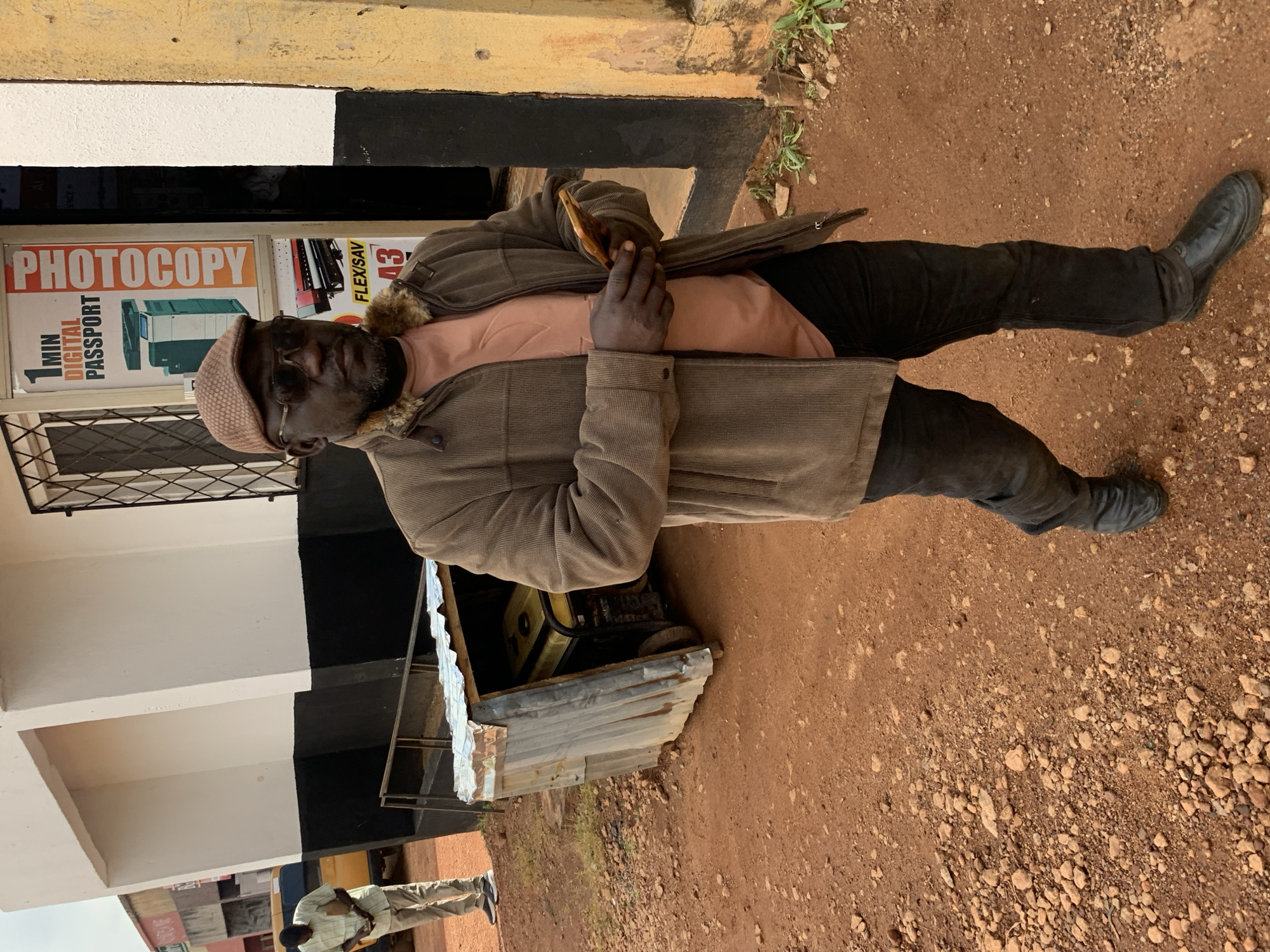A man stands outdoors, facing the camera and looking slightly down. He is wearing a brown corduroy jacket with a fur-lined collar, a light pink t-shirt, dark pants, and black boots. On his head, he wears a patterned cap and dark sunglasses. His right hand is raised, holding a piece of bread to his mouth.
Behind him, a building features a black and white painted facade. A sign in a shop window reads "PHOTOCOPY" in large orange letters, with smaller text below indicating "1 MIN DIGITAL PASSPORT". Other signs with text like "FLEXSAV" are also visible. To the right of the man, part of a generator is visible within a makeshift enclosure made of corrugated metal. The ground is covered in reddish-brown dirt and small stones. The scene appears to be in Gura Top, Nigeria, and the lighting suggests it is daytime. The man's expression is neutral.