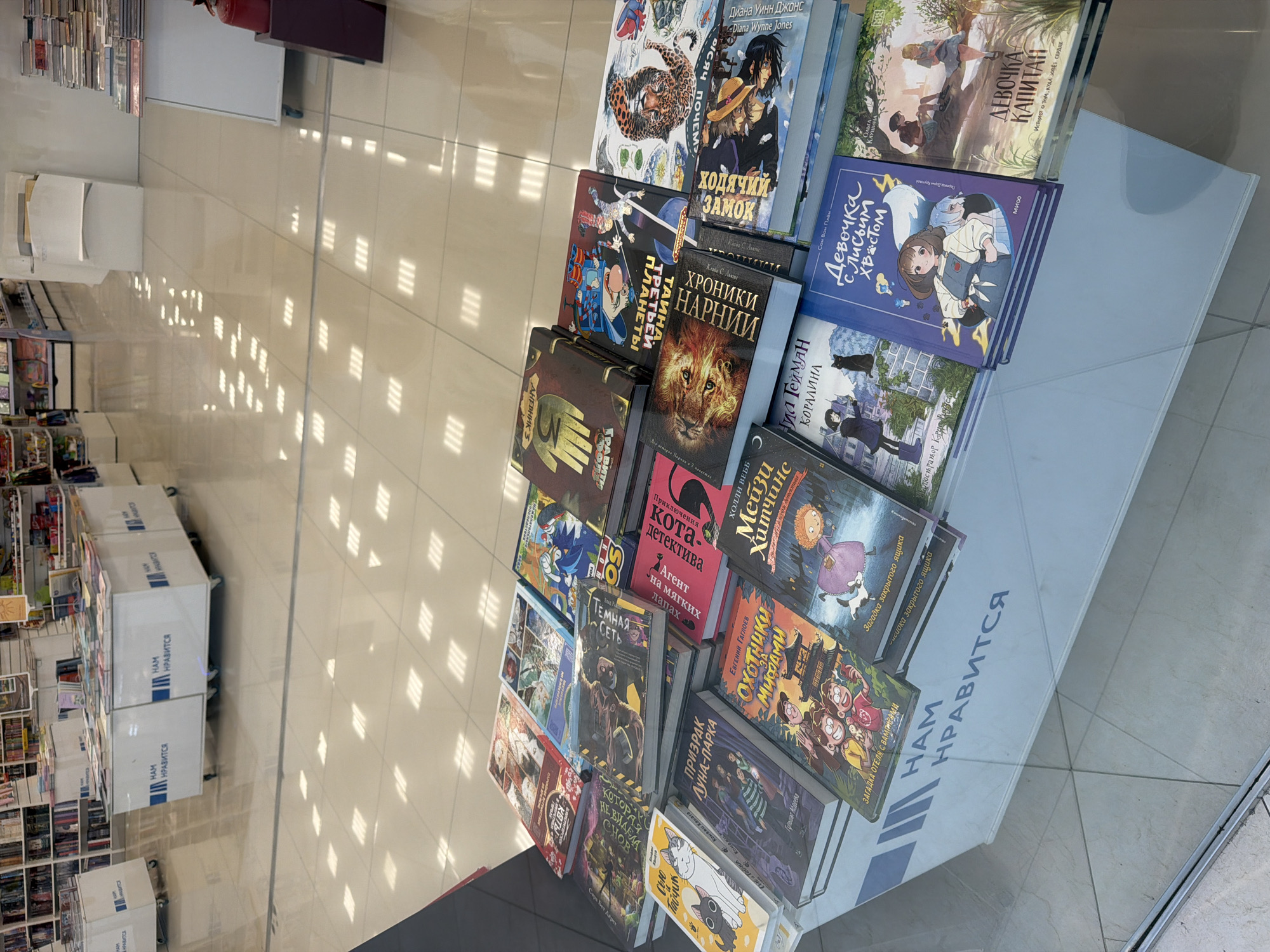 The image shows a display of books in a store in Novoalexandrovka, Russia. The books are arranged on a white shelf or box. The covers of several books are visible, including "ДНЕВНИК 3" (Diary 3), "ХОДЯЧИЙ ЗАМОК" (Howl's Moving Castle), "ХРОНИКИ НАРНИИ" (The Chronicles of Narnia), "КОРАЛИНА" (Coraline), "Девочка с Лисьим Хвостом" (The Girl with a Fox Tail), "ДЕВОЧКА-КАПИТАН" (Girl-Captain), "Приключения кота-детектива" (Adventures of the Cat-Detective), "ОХОТНИКИ ЗА МИФАМИ" (Myth Hunters), "Мейзи Хитчинс", "Призрак Луна-Парка" (The Ghost of the Moon Park). The phrases "НАМ НРАВИТСЯ" (We Like It) are printed on the sides of the display. The floor is tiled. Other shelves of books are visible in the background.