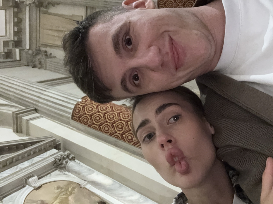 Two individuals, a female and a male, are captured in a selfie-style close-up photograph. The female, positioned on the left, has fair skin, dark eyebrows, and puckers her lips in a "duck face" expression. Her head is tilted and rests against the male's shoulder. She wears a plaid garment partially visible and a ribbed, neutral-toned knit sweater. The male, on the right, has short, dark hair and fair skin. He displays a slight closed-mouth smile with wide eyes directed at the camera. He wears a white t-shirt under a dark, possibly olive or brown, cardigan.
The background reveals elaborate interior architecture, characteristic of a historical building in Venice, Italy. Visible elements include a white marble fluted column with an ornate capital, decorative cornices, and a segment of a fresco depicting a figure in a curved architectural recess. A patterned textile, displaying a repeating geometric or curvilinear design in reddish-brown and cream tones, is visible as a wall covering or drapery behind the subjects. The scene is adequately lit, illuminating the architectural details.