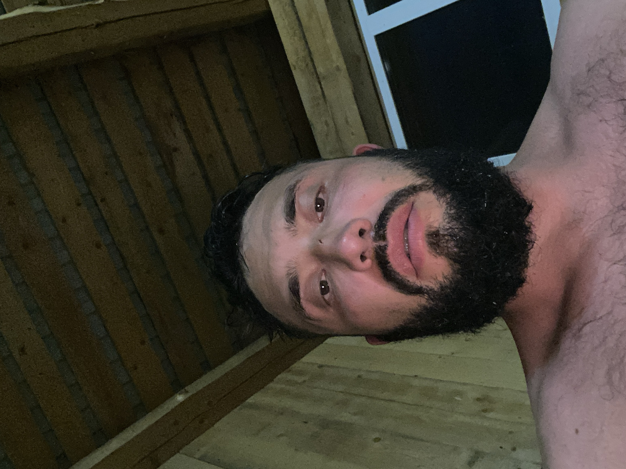 The image is a close-up, low-angle selfie of a man with a beard, taken from the chest up. He appears to be indoors, in what looks like a rustic structure. The background shows wooden walls and a wooden ceiling with exposed beams. The lighting is dim and warm. The man is looking directly at the camera, with his mouth slightly open. There is a window visible in the background to the right of the man. The environment suggests a rural or possibly unfinished building. The specific location within Zhigalki, Russia cannot be determined from the visual information alone.