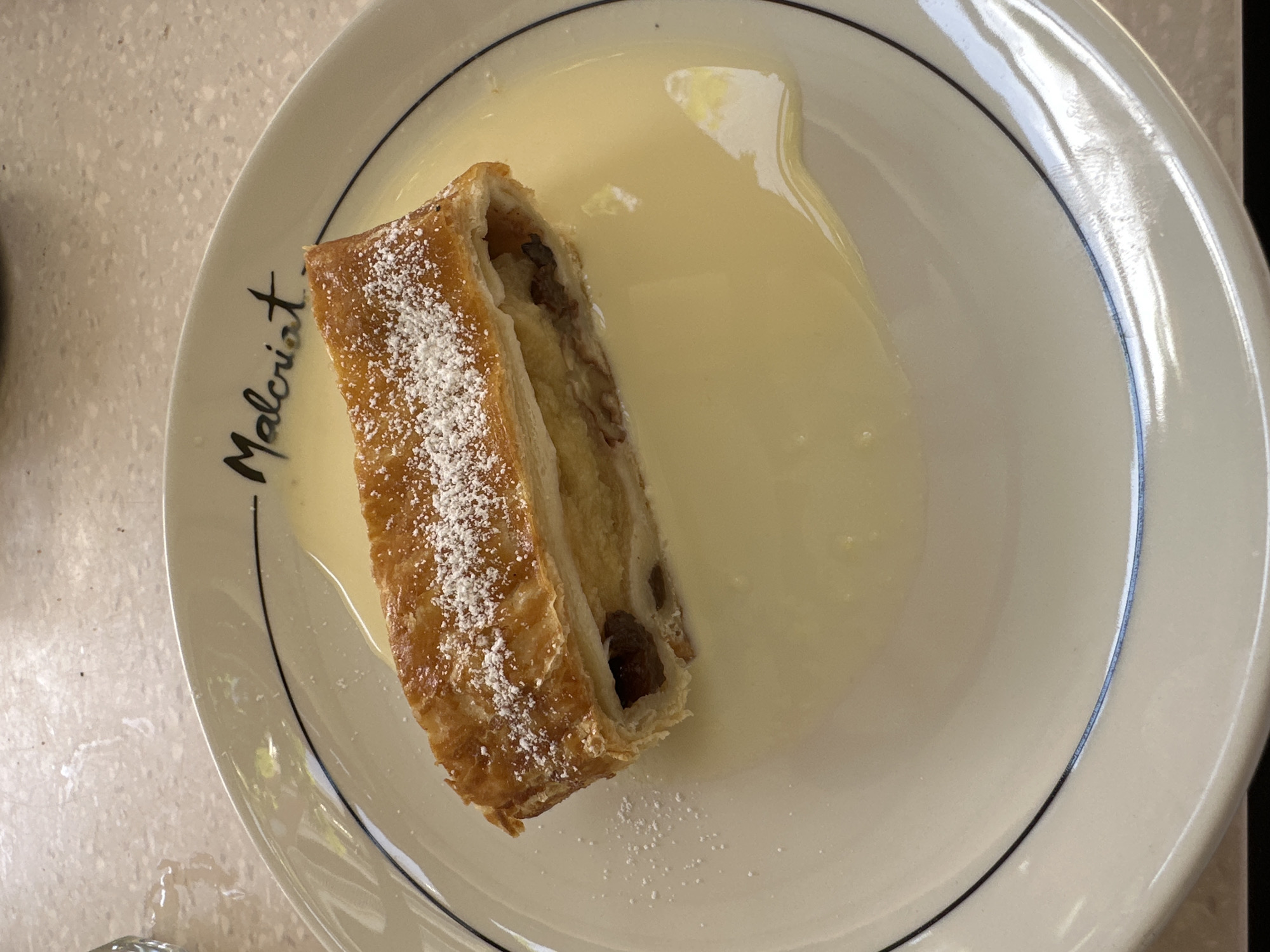 The image shows a plate of apple strudel covered in powdered sugar, sitting in a pool of light yellow sauce. The plate is white with a black rim and has the word "Malcriat" printed on the edge. The food appears to be served indoors, on a table with a light-colored surface. While I can't pinpoint the exact location in Sant Feliu de Guíxols without additional clues, the presence of "Malcriat" on the plate suggests that it was likely served at or associated with a restaurant or establishment of that name in the city.