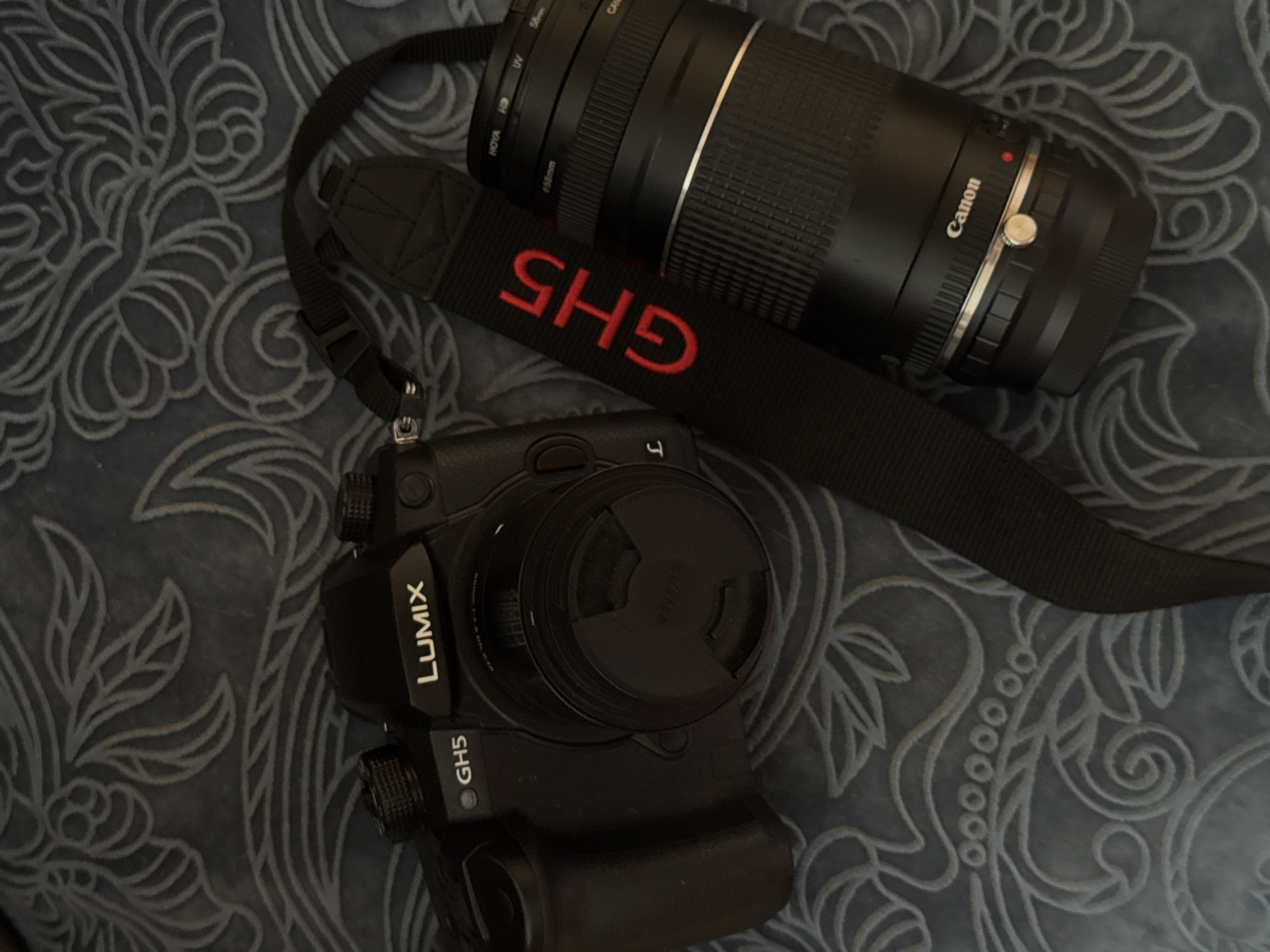 The image shows a Lumix GH5 camera with a lens cap, a Canon lens, and a camera strap with "GH5" embroidered in red. The objects are arranged on a patterned, possibly fabric surface. The location of the photograph is not apparent from the image and no identifying information related to Izhevsk, Russia is present.
