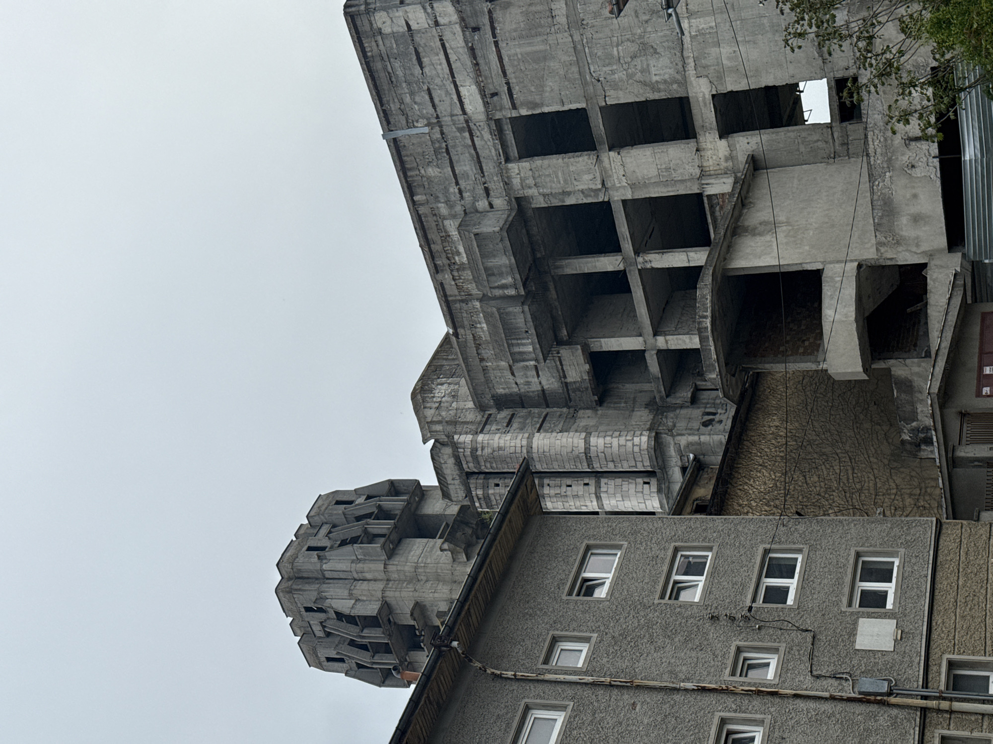 The image depicts an upward shot of several brutalist concrete buildings in Shumen, Bulgaria. The sky is overcast. A taller building under construction dominates the view with its exposed concrete structure, visible open floors, and grid-like pattern. Adjacent to this structure is a shorter, older building with rows of rectangular windows. The facade of this building is textured. Some wires run along its surface. In the upper right corner, there are some leaves and part of a corrugated metal fence.
