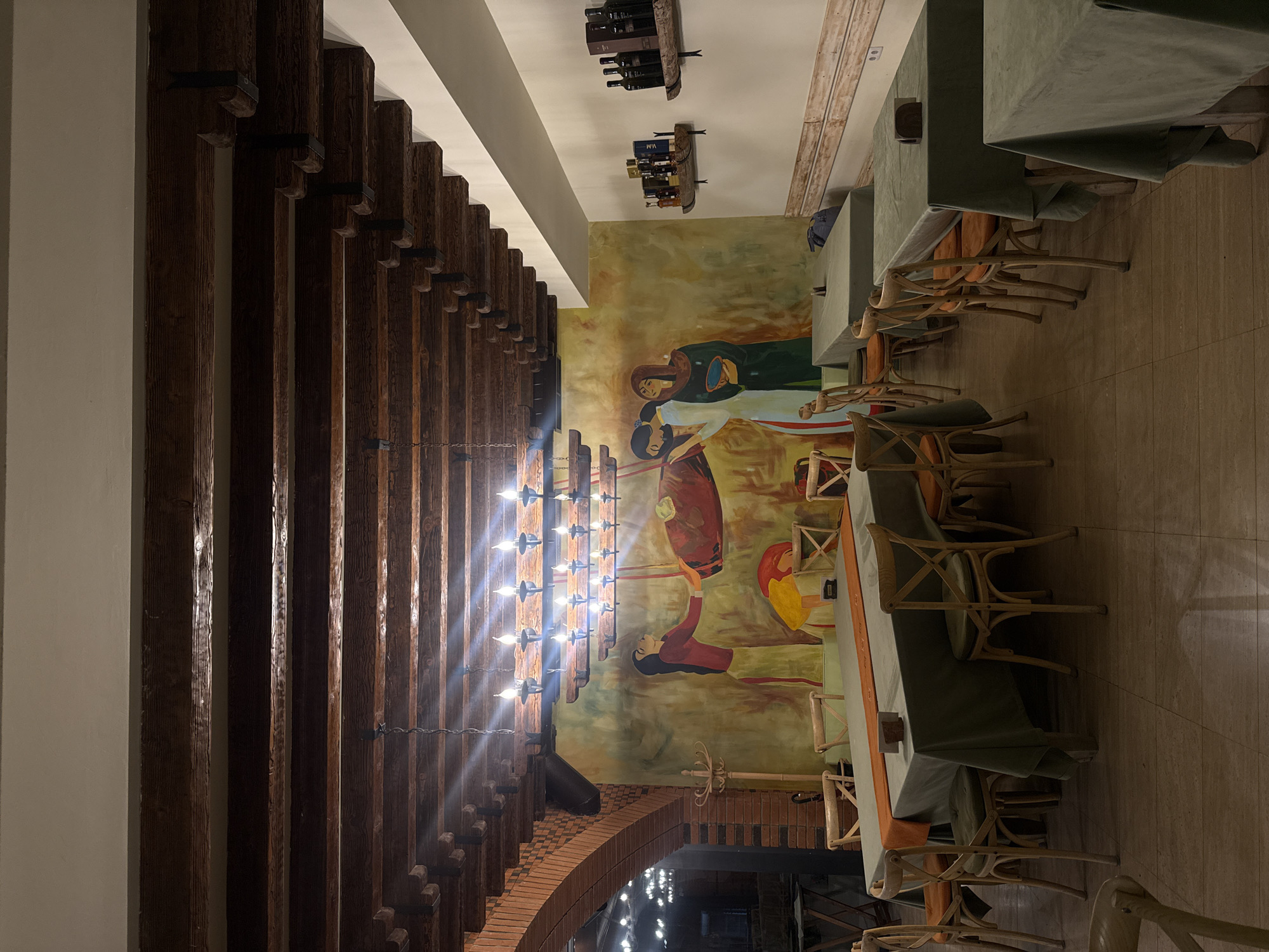 The image depicts the interior of a restaurant in Sasunik, Armenia. The ceiling features exposed wooden beams and a chandelier with multiple lights. The walls are adorned with a mural of figures and a large round object. Several tables, covered with green tablecloths and accented with orange runners, are set up for dining, each surrounded by wooden chairs. On the right wall, two wooden shelves display bottles of wine and other items. An arched brick structure is visible on the left side of the image, and a coat rack stands near the mural. The floor is tiled. No people are visible in the scene.