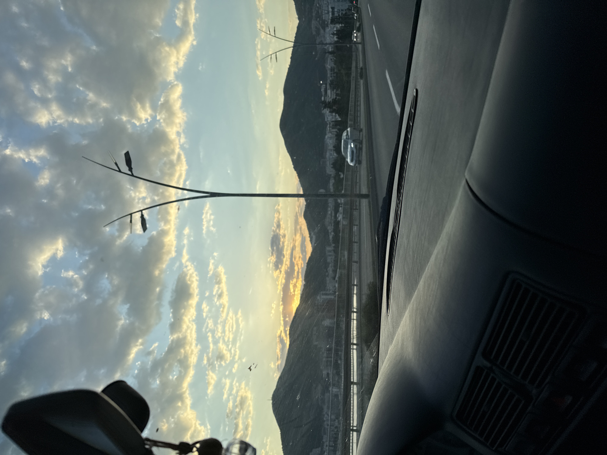 The image shows a view from inside a car, looking out onto a highway in Budva, Montenegro. The sky is filled with light blue and gold-tinged clouds, suggesting either sunrise or sunset. In the middle ground, there is a mountainous landscape dotted with buildings, indicating urban development. A distinctive lamppost with minimalist, branch-like arms and modern light fixtures is visible on the roadside. The road features multiple lanes and a median barrier. The car's dashboard and interior elements are visible in the foreground, including the rearview mirror and air vents. A glimpse of another vehicle can be seen on the highway. Overall, the scene depicts a journey along a coastal highway in Budva, likely at dawn or dusk.