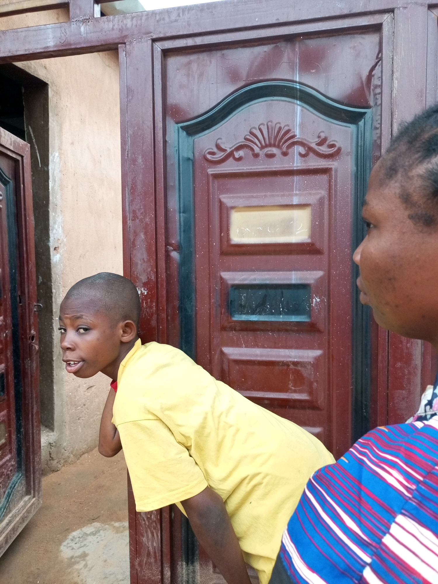 In Tempure, Nigeria, a child with a shaved head and a bright yellow shirt leans out from behind a burgundy metal door, looking to the left with an open mouth. Their left arm is bent, and their left hand rests on the door frame. A woman, partially visible on the right side of the frame, wears a colorful patterned shirt of blue, red, and white stripes. The background consists of a textured beige wall and more of the burgundy metal door, which has decorative molding and rectangular insets.