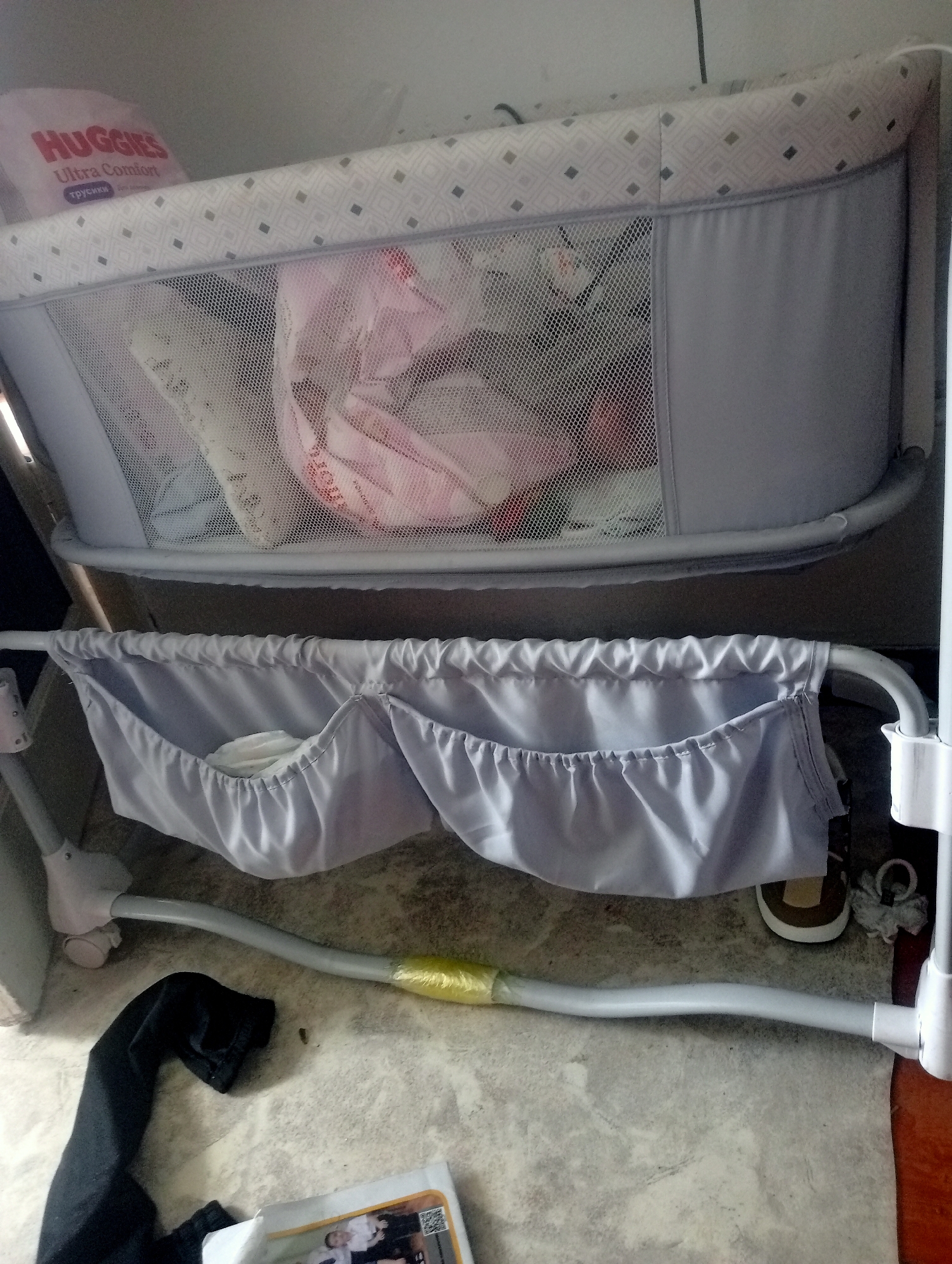 An interior shot captures a baby bassinet with a mesh siding, revealing baby items within. Attached to the lower frame of the bassinet is a grey fabric organizer with multiple pouches, one of which contains a stack of white baby wipes. The bassinet sits on a floor with a patterned tiled surface. To the left, a black garment lies on the floor. In the lower right corner, a portion of a brown shoe is visible, along with a white circular object. The image does not provide enough information to determine the location within the city of Aksu, Kazakhstan.
