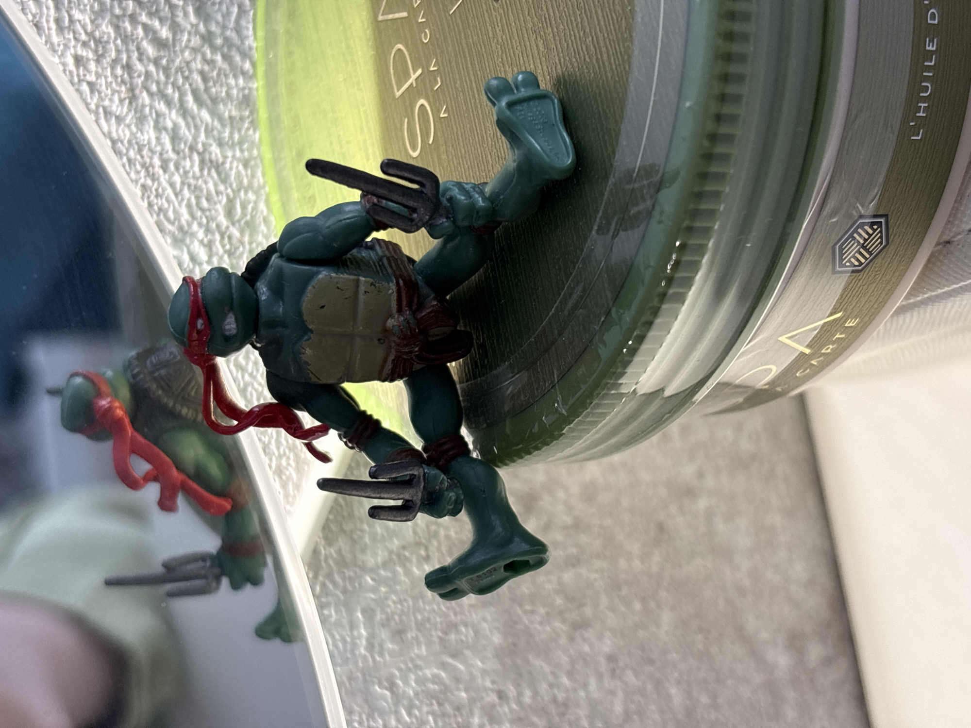 A Teenage Mutant Ninja Turtle action figure, specifically Raphael with a red mask and sai weapons, is positioned on top of a green jar of hair product. The jar has the word "SPA" visible on its label along with other unreadable words. A partial reflection of the same action figure is visible on a nearby reflective surface. The background includes textured beige walls and an unknown item in the background. The image provides no evidence or reference to the city of Izhevsk, Russia.