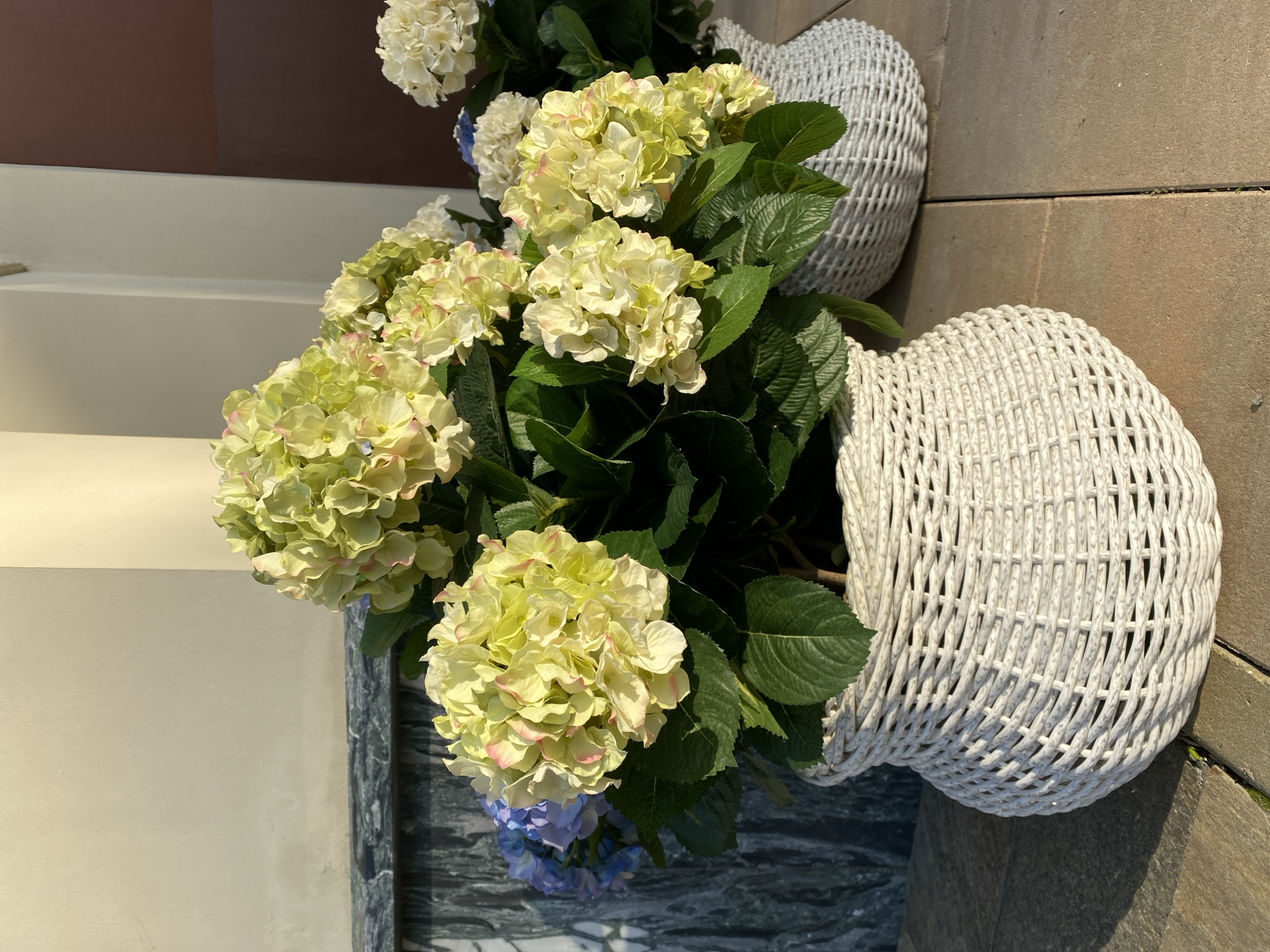The image displays two white, woven-patterned vases filled with flowering plants, including hydrangeas with green, white, and hints of pink/purple petals, and green leaves. The location appears to be outdoors, possibly in a garden or patio setting, given the presence of stone flooring and building elements. There is no verifiable information that it is in Yekaterinburg, Russia. There are no people present in the image.