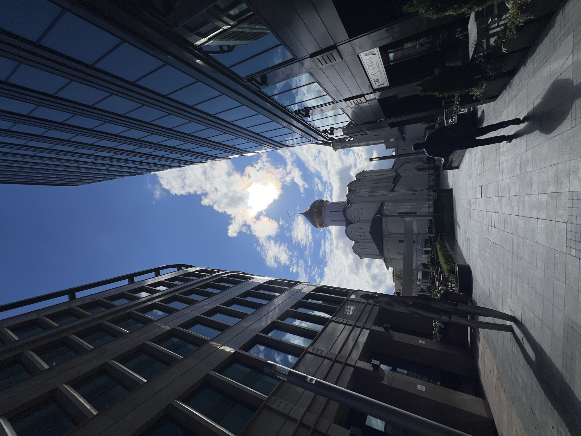 The image depicts a street view in Moscow, Russia. On the right, a modern glass-facade building reflects the sky. On the left, a building with a grid-like window pattern stands. Between the buildings, the sky is visible with the sun obscured by clouds. The gold dome of a Russian Orthodox church appears in the distance. On the right sidewalk, a person is walking away from the camera, casting a shadow. A sign for "TOKYO GRILL" is visible on the building to the right. The street is paved with rectangular stone slabs.