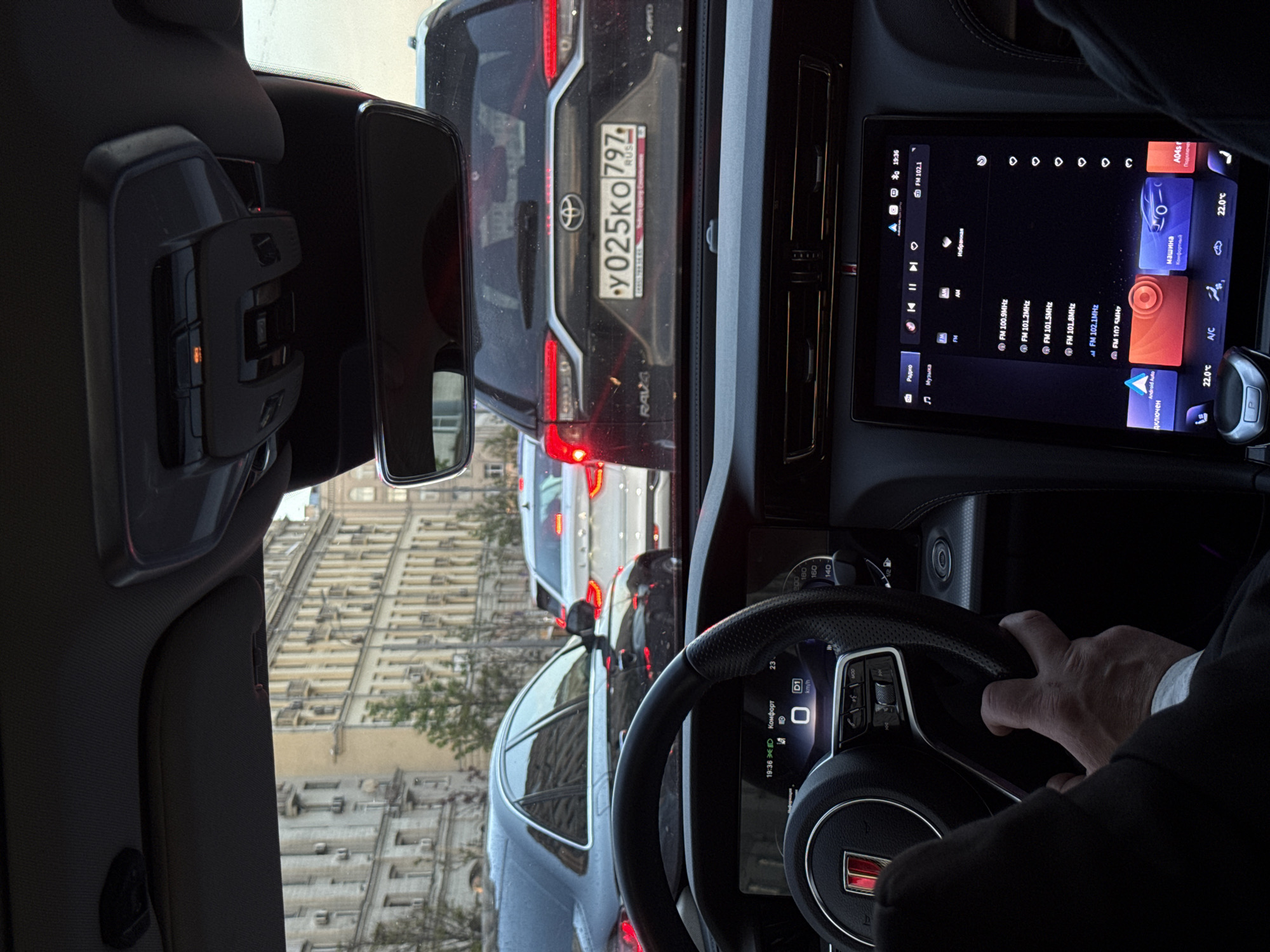 The view from inside a car shows the car is stationary in traffic. The license plate of the vehicle in front is y025KO 797, a Russian license plate. The car in question is a Toyota Rav4. The infotainment system shows it is playing radio, FM 102.1. A hand is visible on the steering wheel. The building seen through the front windshield is typical of Moscow architecture. The image was taken at 19:36.
